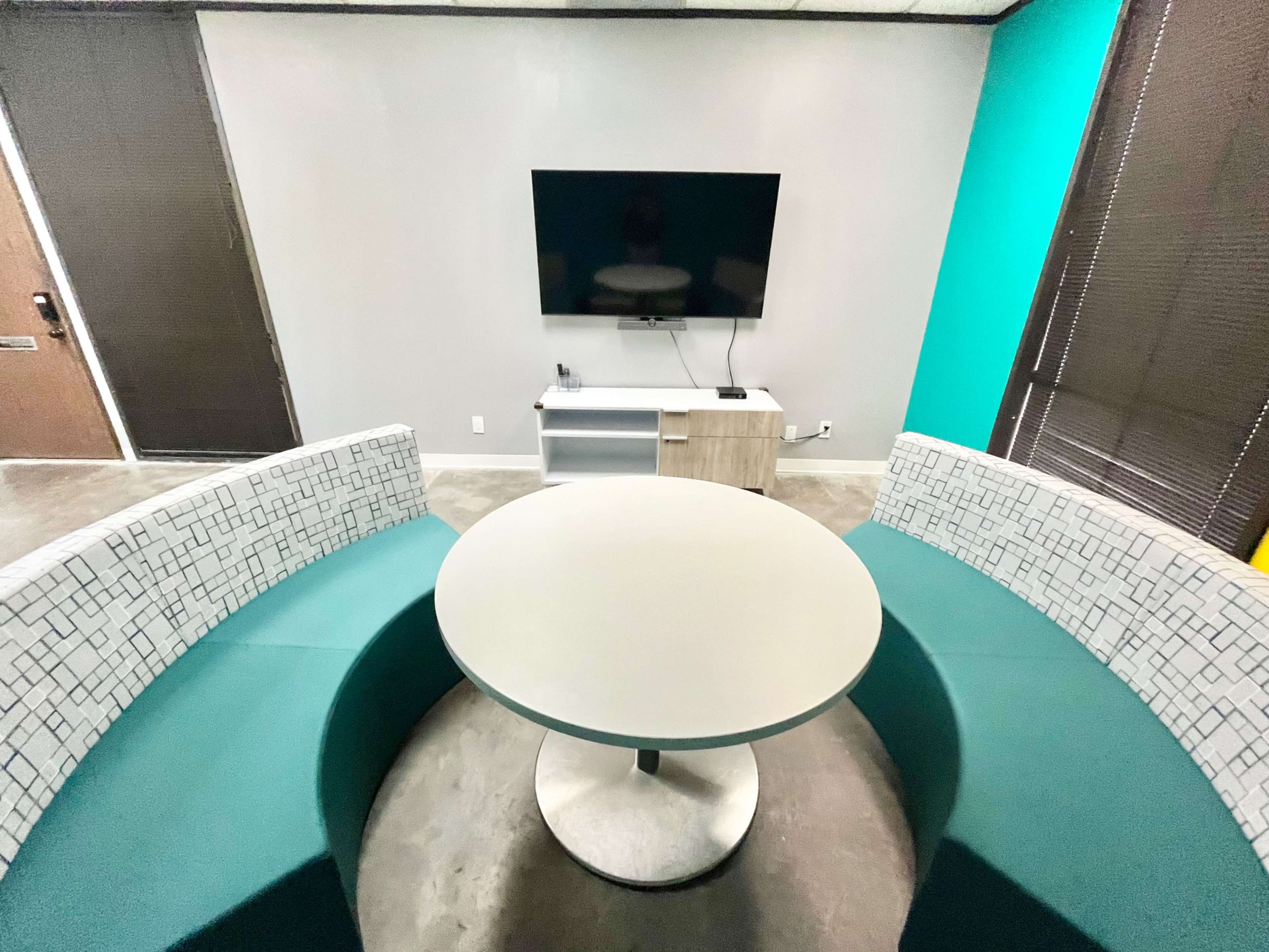 The image shows a modern seating area with a circular table and two curved benches arranged around it, facing a wall-mounted television.