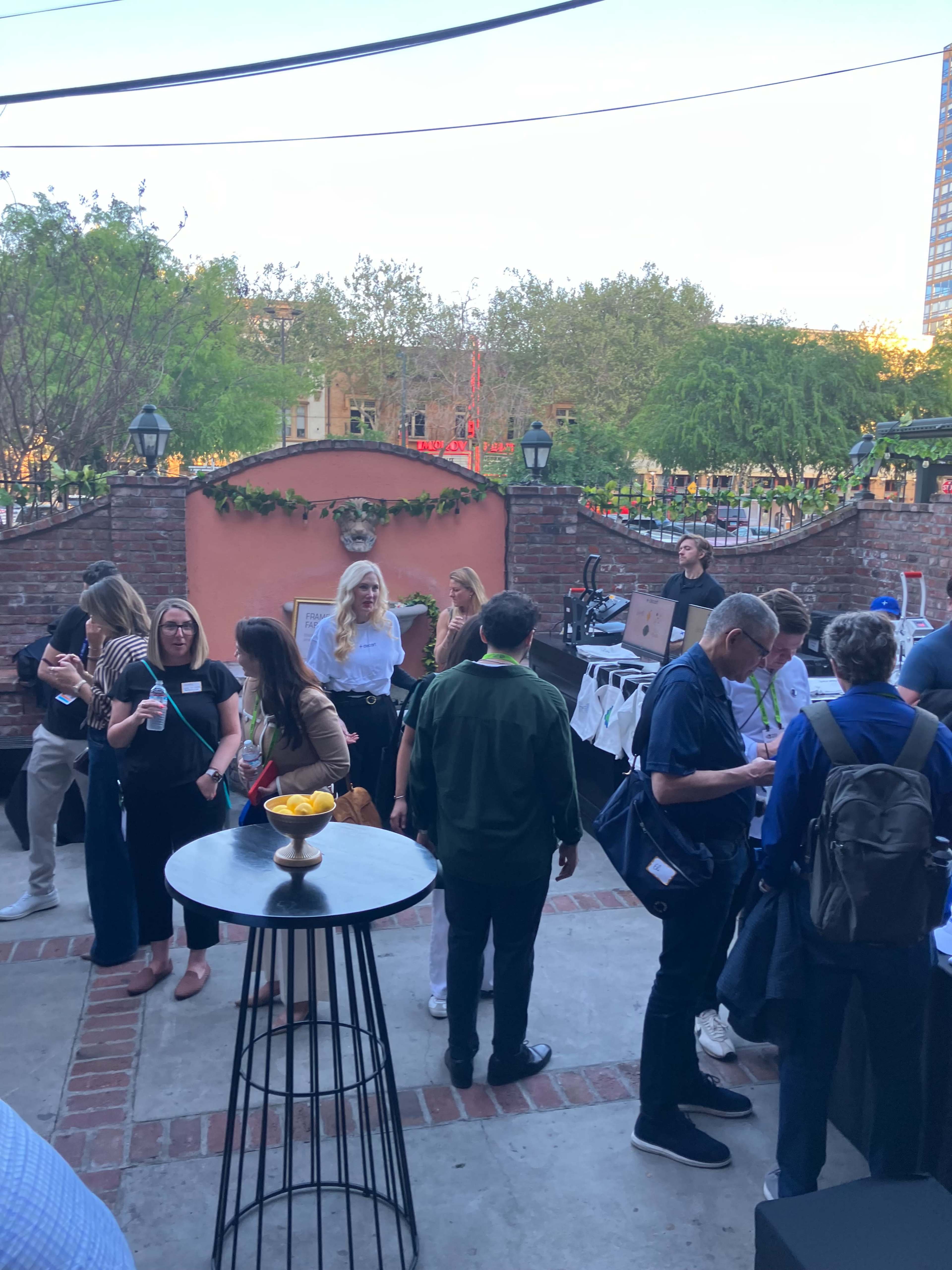 A group of people socialize at a gathering in an outdoor patio area, surrounded by greenery and illuminated by soft lighting.