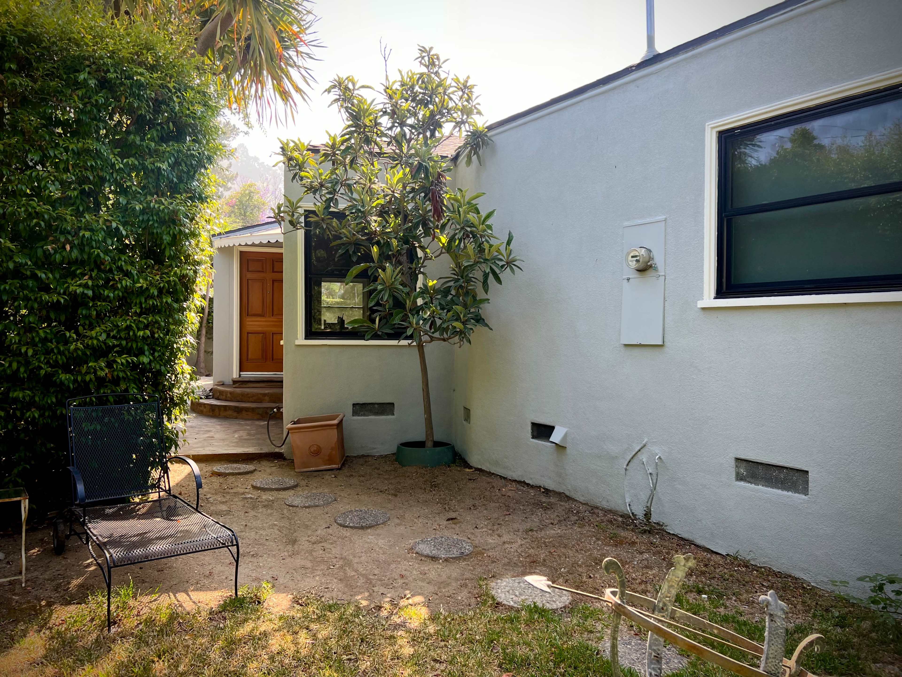 A small, landscaped backyard features a tree, a pathway of stepping stones, and a chair beside a white wall with a window.