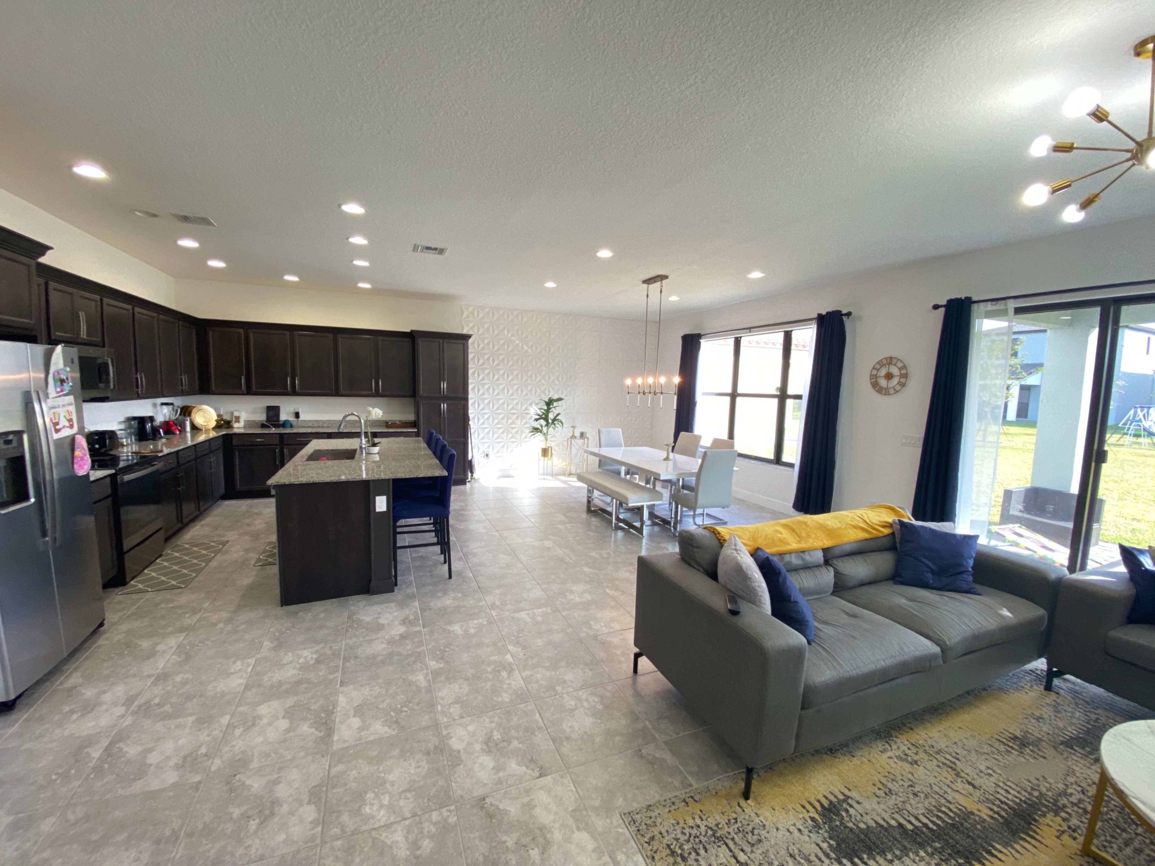 A modern kitchen and dining area with dark wooden cabinetry, stainless steel appliances, and a spacious layout featuring a gray sofa and a dining table with white chairs.