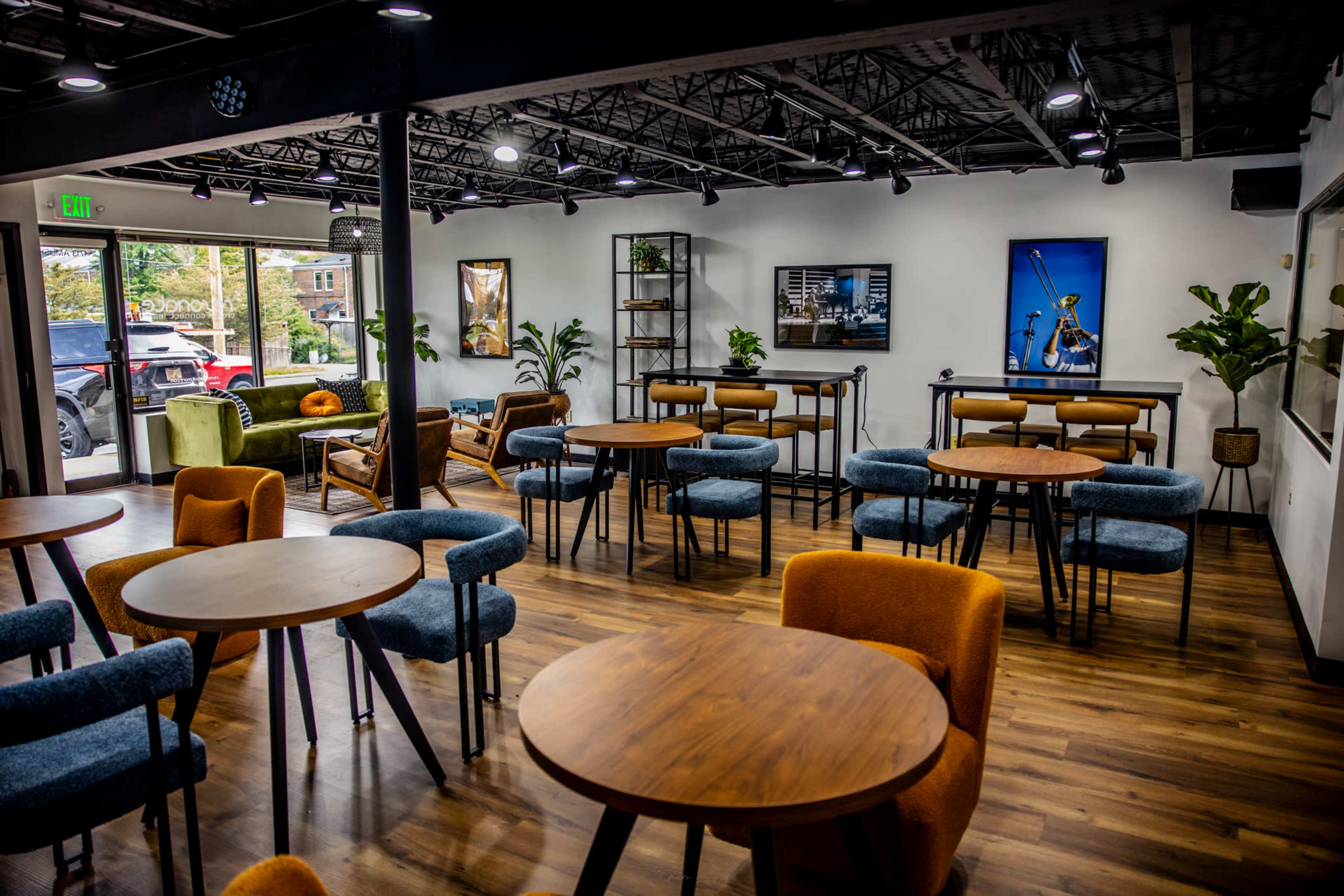 A modern cafe interior with wooden tables and chairs arranged around a spacious area featuring plants, artwork on the walls, and large windows.