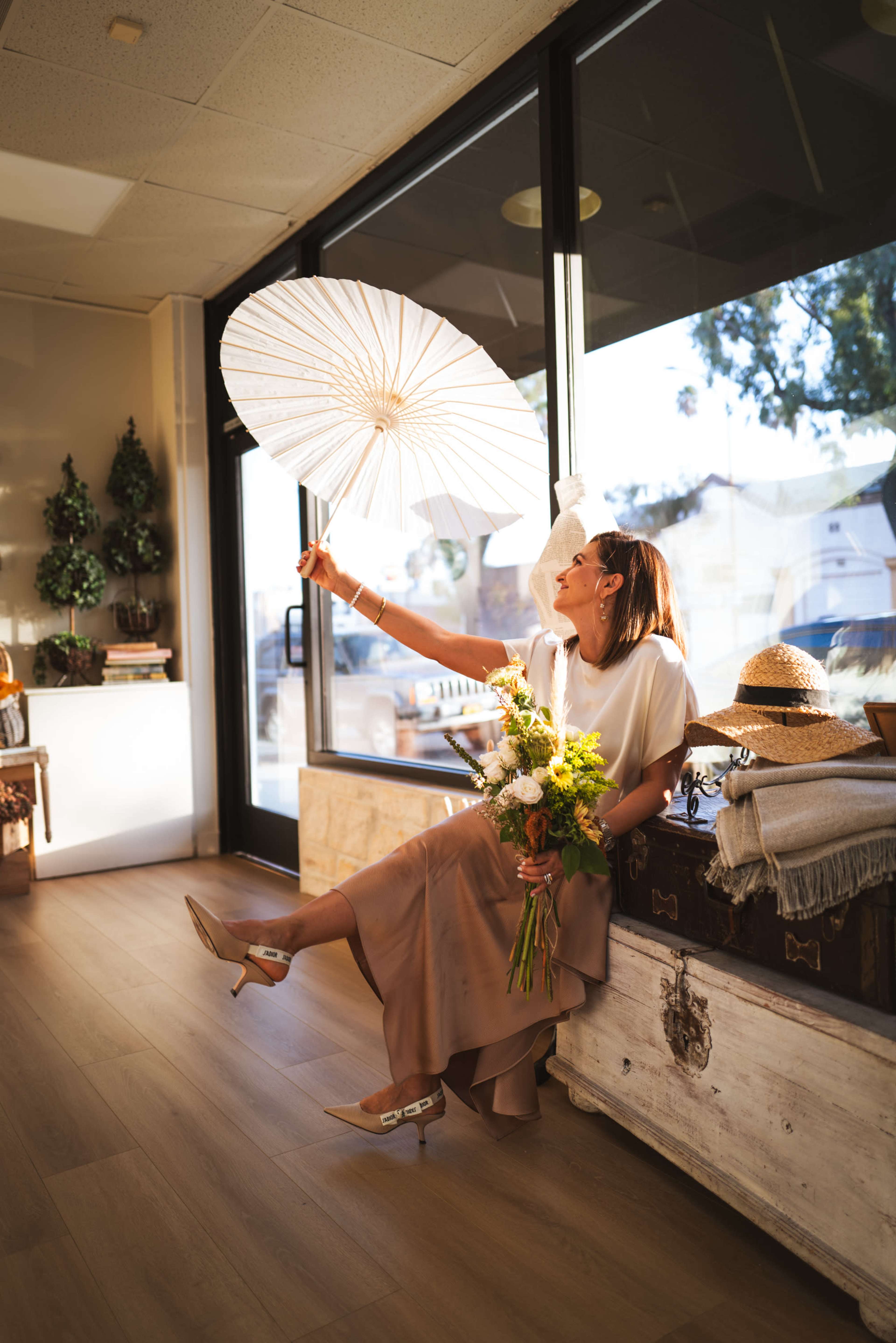 A woman holds a white parasol and a bouquet of flowers while sitting on a chest in a sunlit room with large windows.