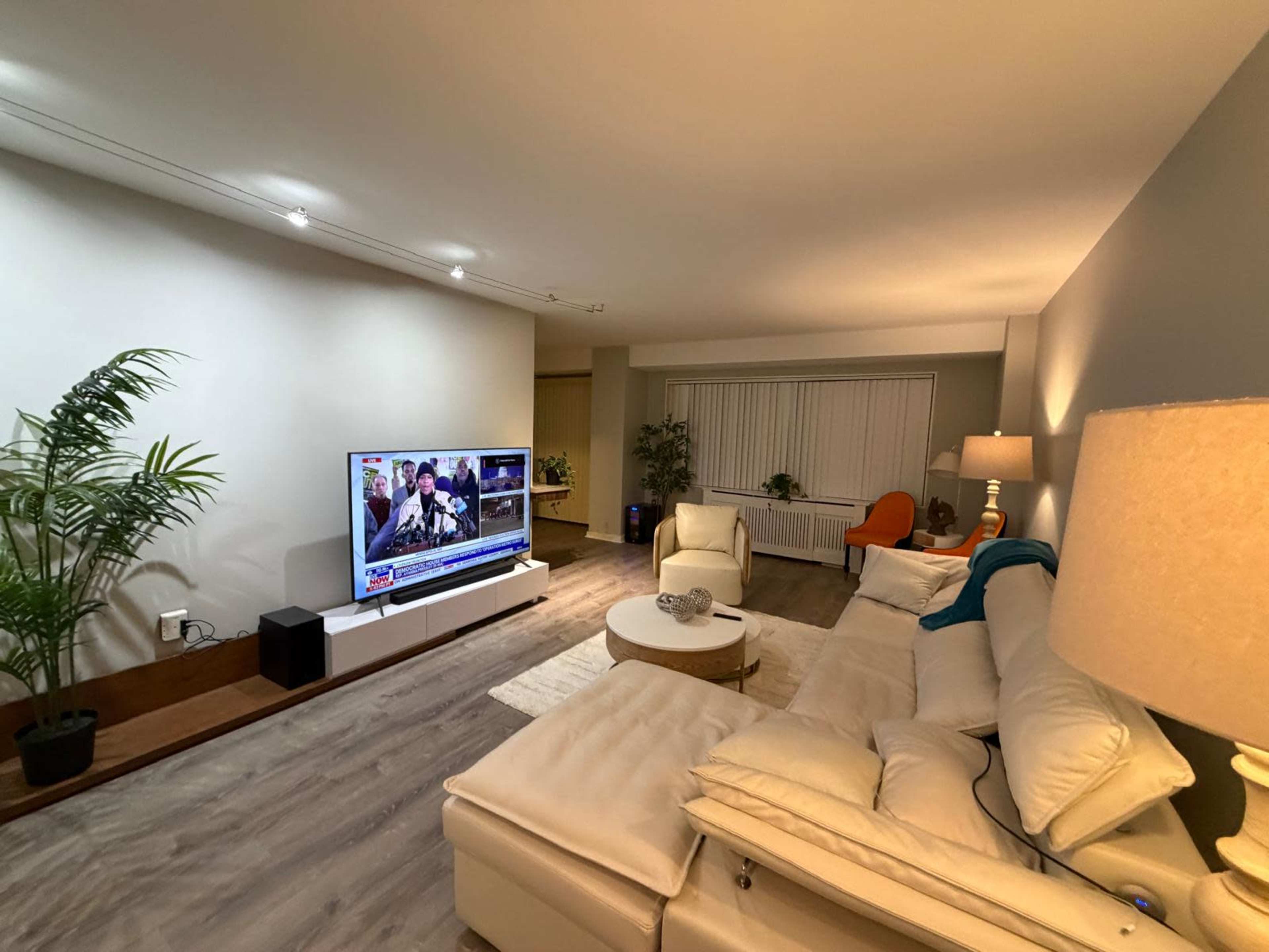 The image shows a modern living room with a large television, a white sectional sofa, a round coffee table, and a plant in a corner, illuminated by soft lighting.