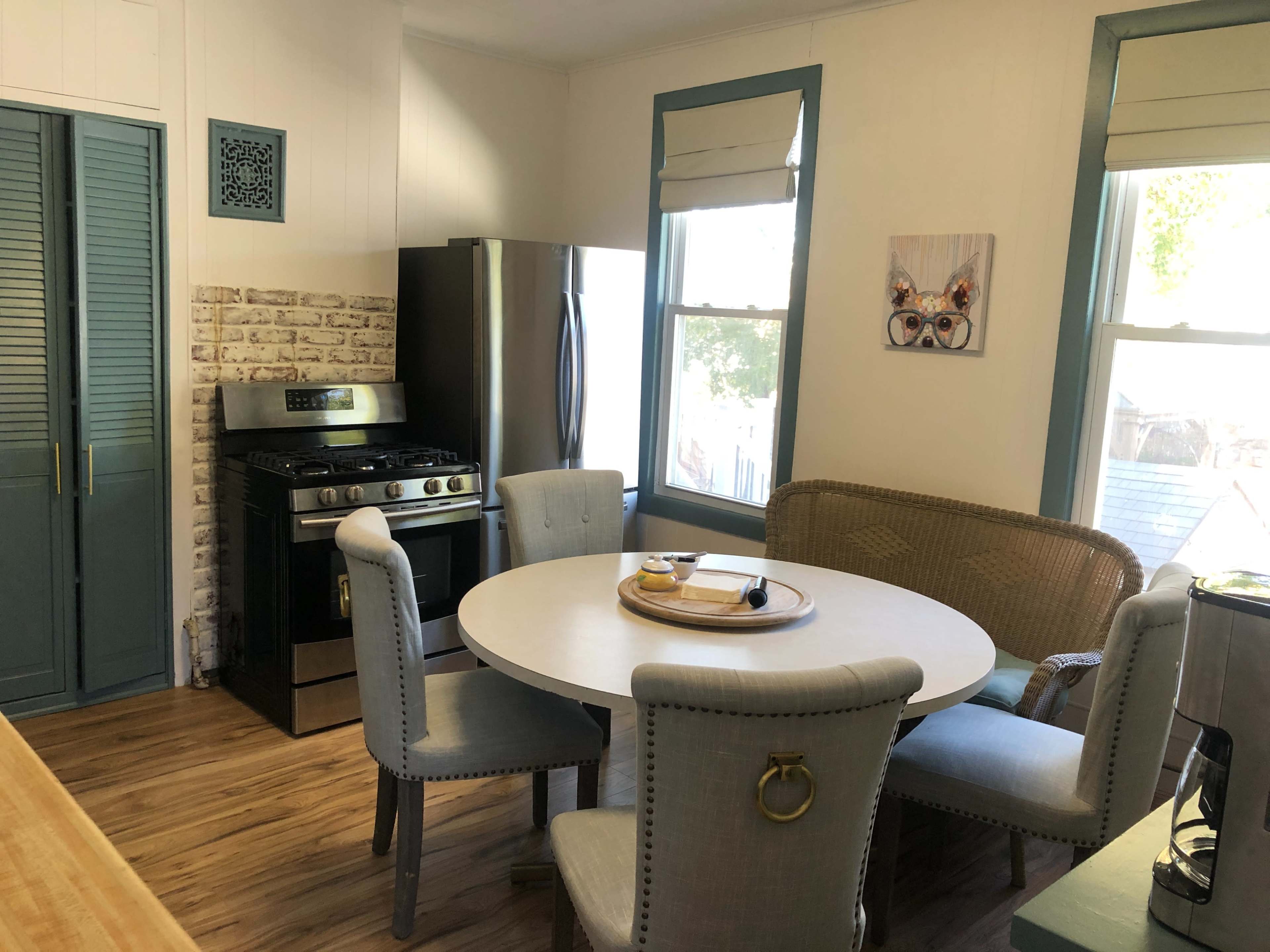 The image shows a kitchen with a round table surrounded by upholstered chairs, a stainless steel refrigerator, and a gas stove against a wall with brick accents.