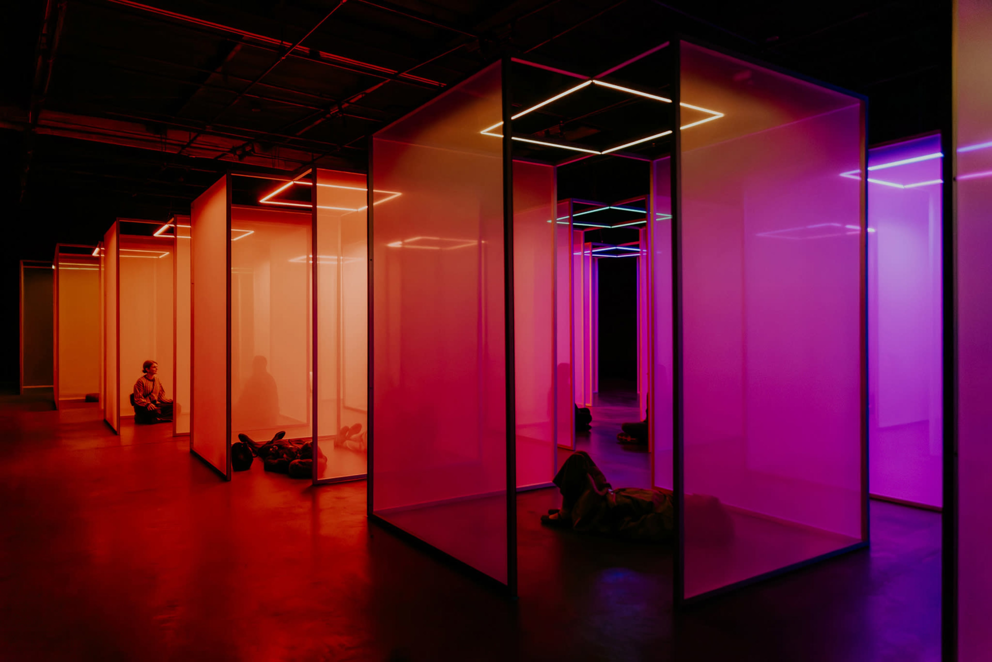 The image depicts a spacious interior lined with illuminated, transparent partitions in shades of pink and orange, with people seated or lying within some of the sections.