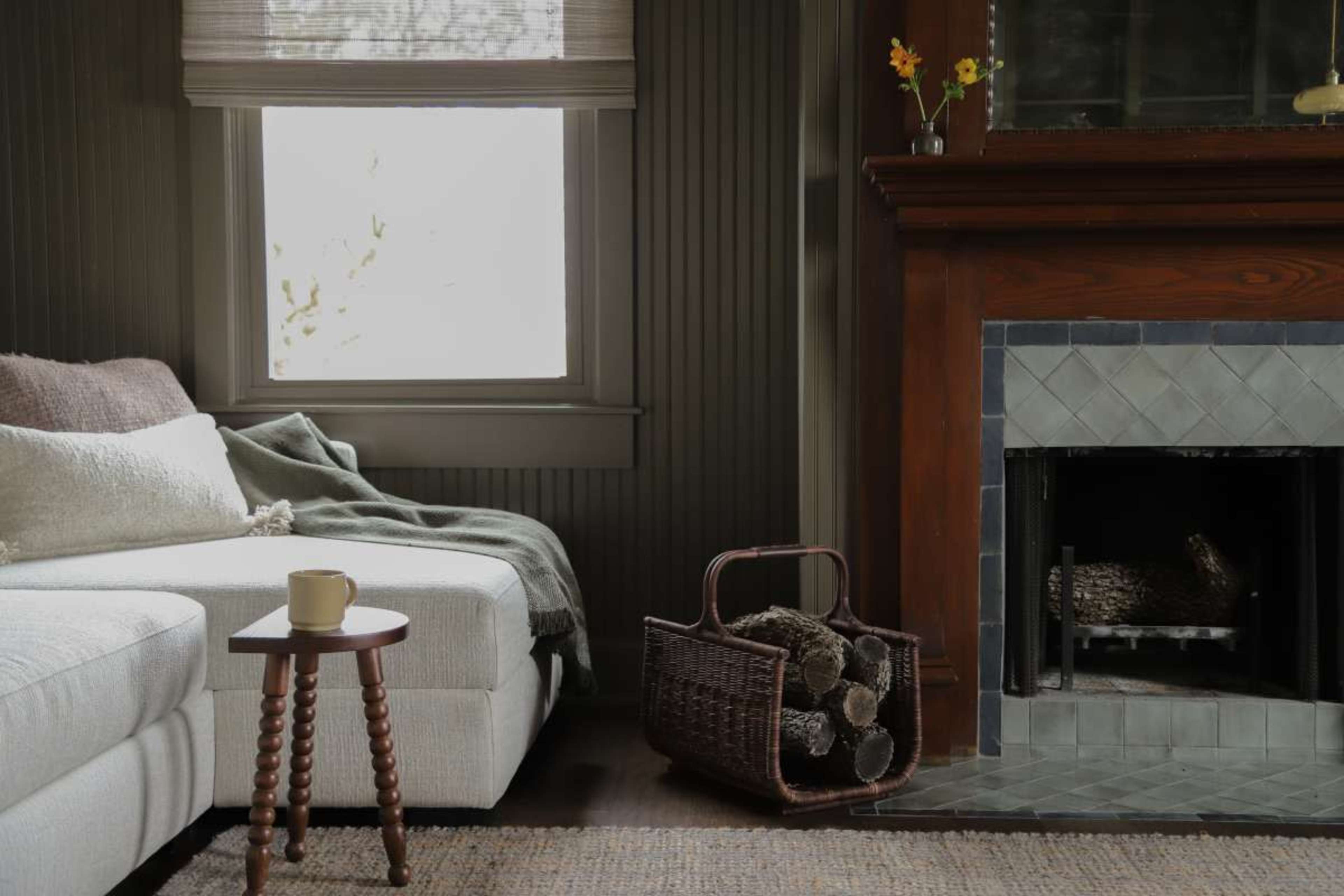 A cozy living room features a white sectional sofa, a small wooden side table with a cup, a woven basket of logs, and a fireplace with a stone surround.