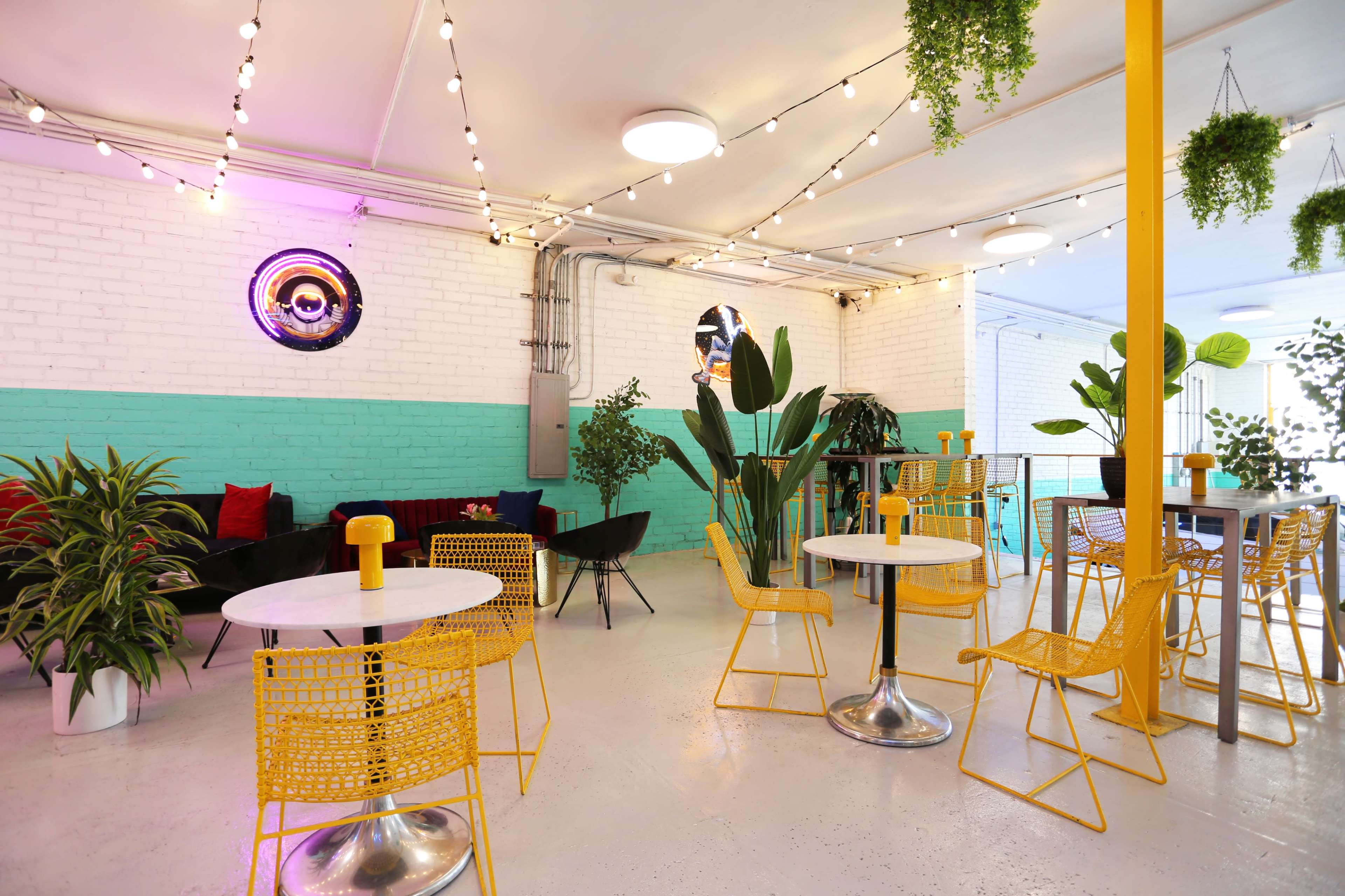 A brightly lit café with colorful seating, greenery, and string lights hanging from the ceiling.