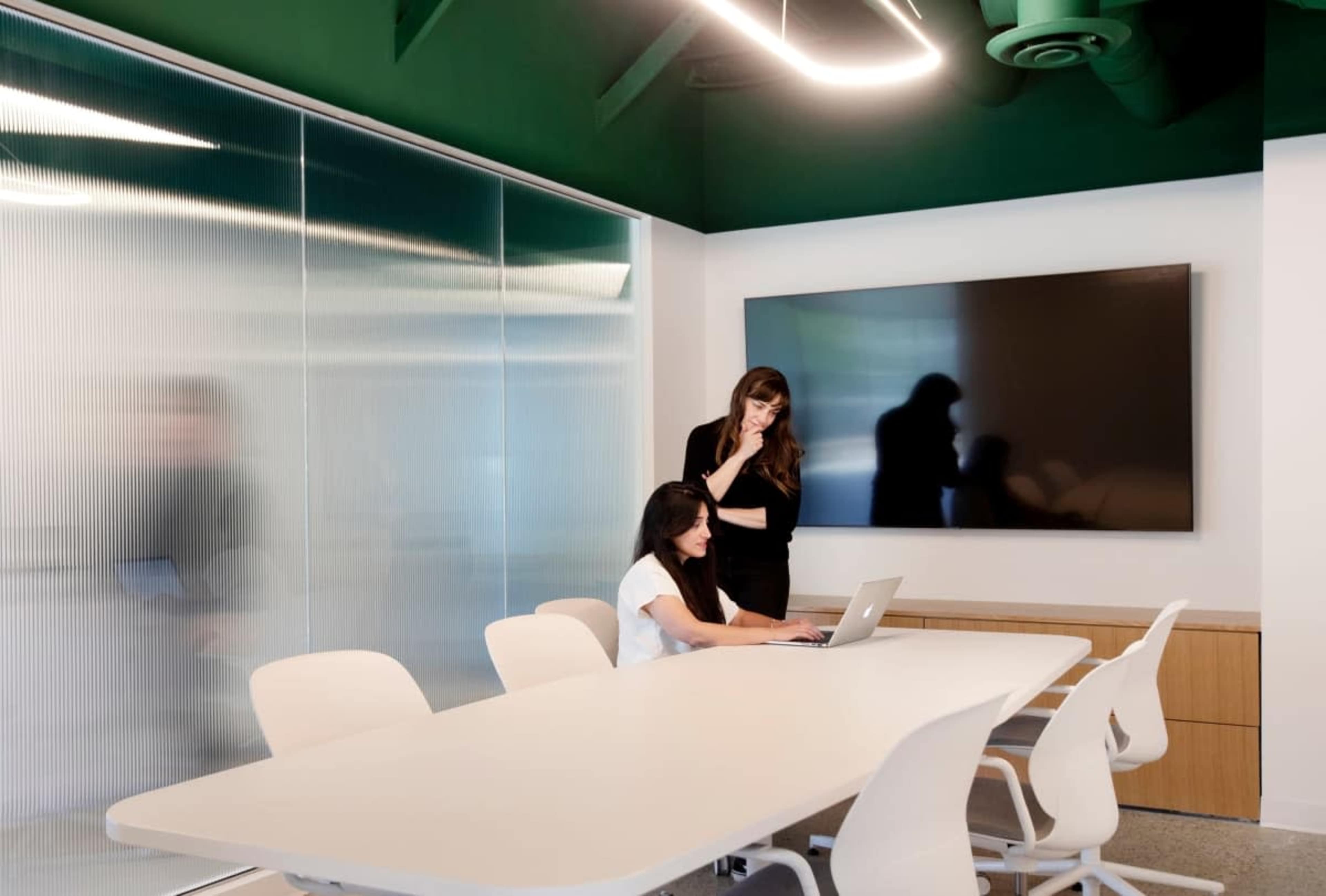 Two women collaborate in a modern conference room, one seated at a laptop and the other standing nearby, while a large screen displays on the wall.