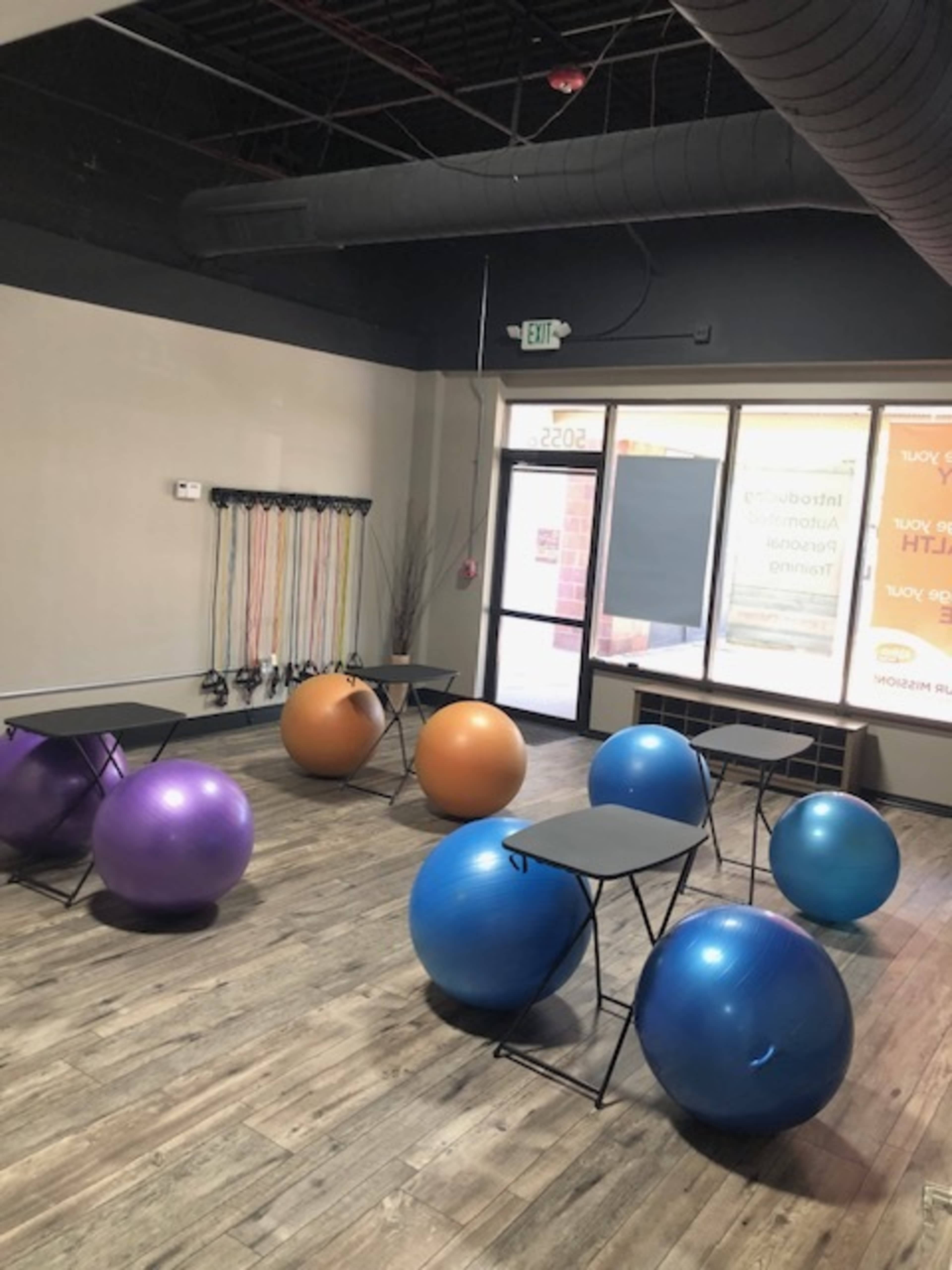The image shows a fitness studio with two tables and several large exercise balls in different colors, placed on a wooden floor.
