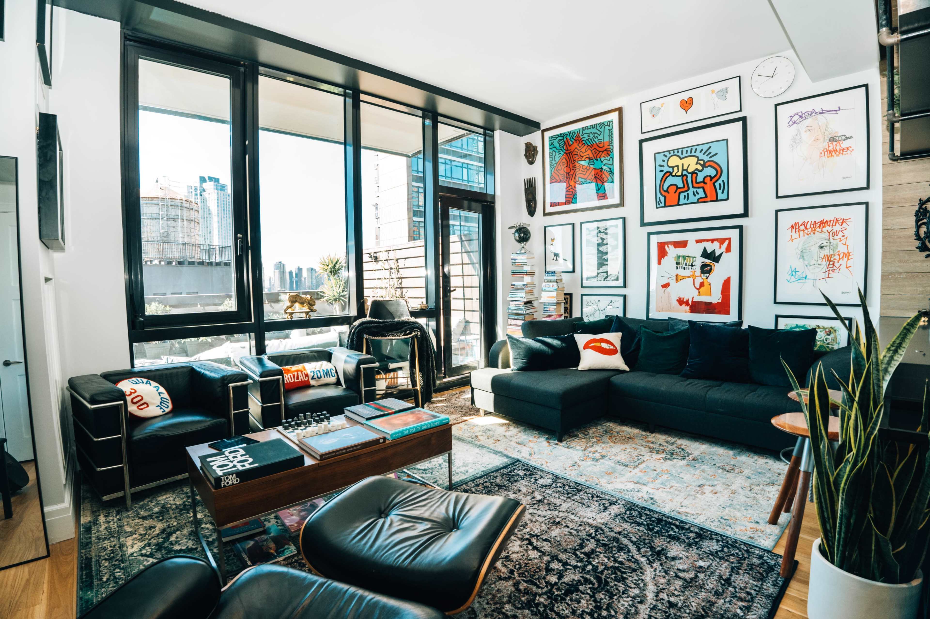 The image shows a modern living room with large windows, a black sectional sofa, various framed artwork on the walls, and a wooden coffee table on a patterned rug.