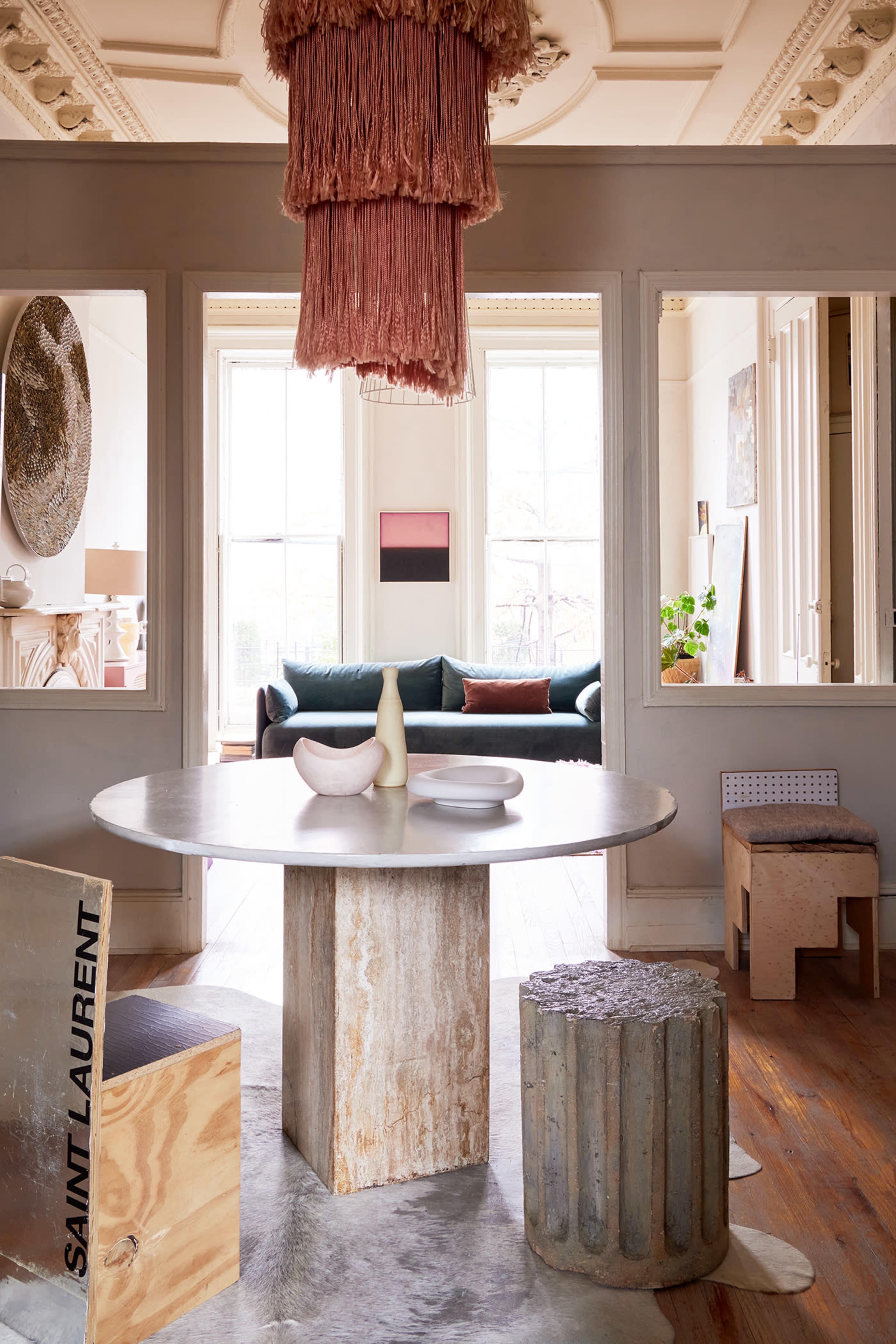 A modern dining area features a round table with a concrete pedestal, surrounded by unique chairs, and illuminated by a pink fringe chandelier.