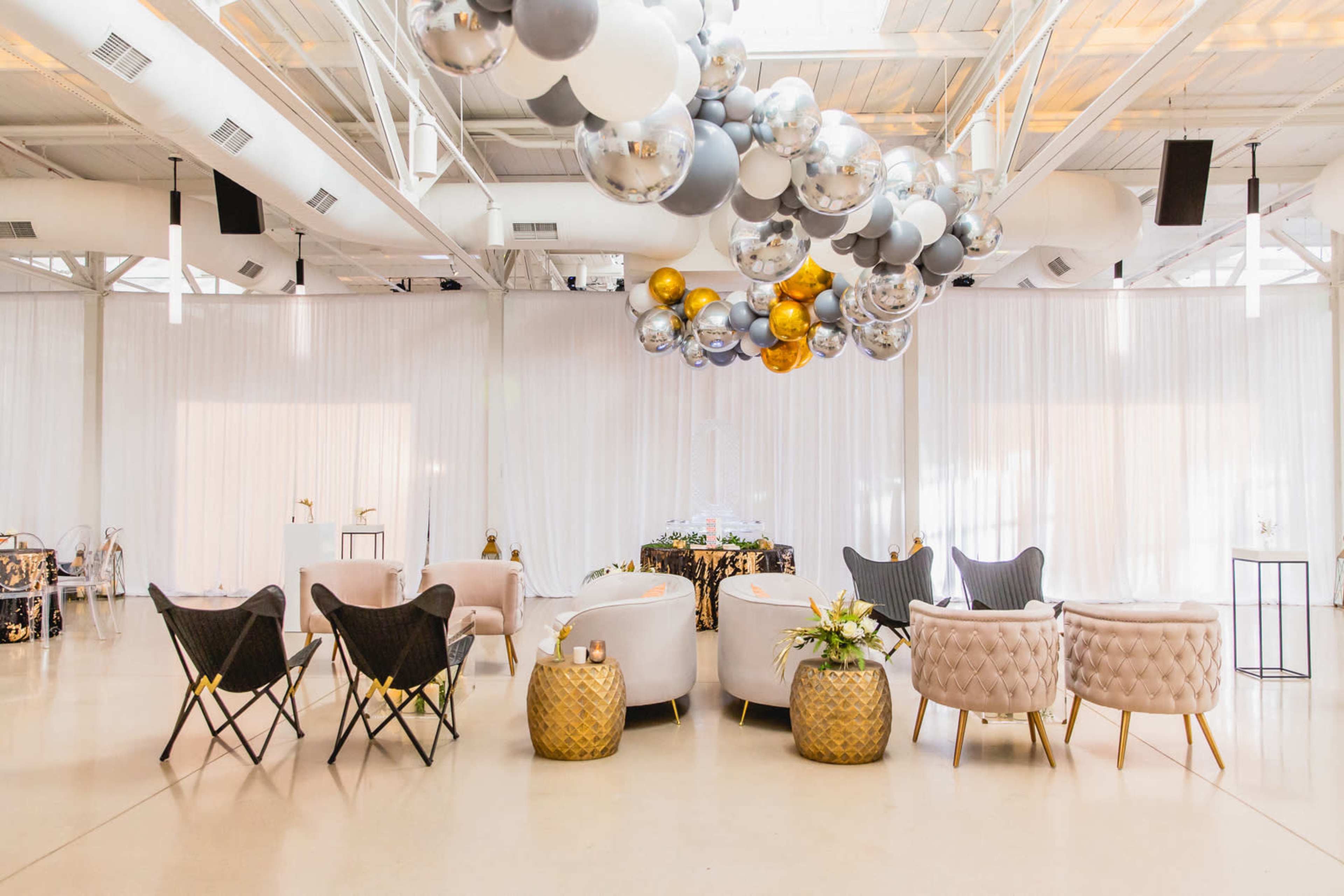 A decorated indoor event space with a cluster of balloons in silver, gold, and black hanging from the ceiling, accompanied by elegantly arranged seating and tables.