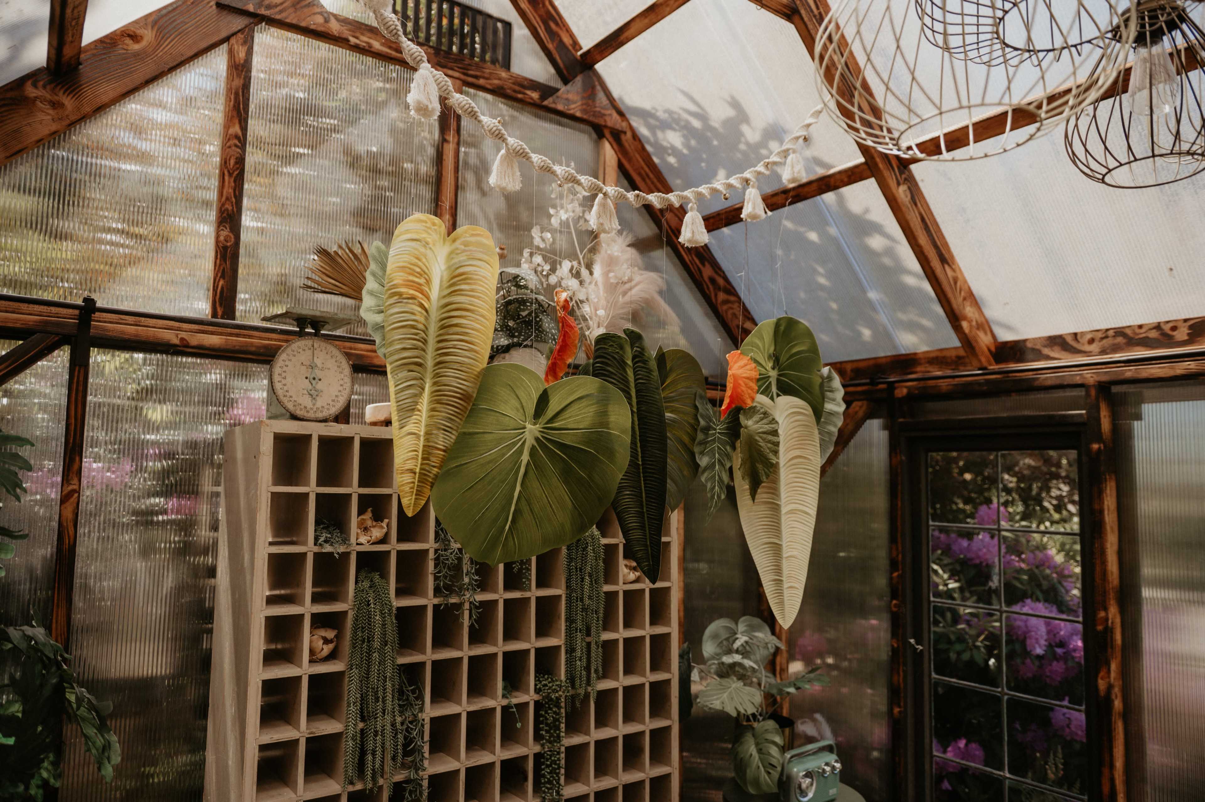 The image shows a greenhouse interior with large, leaf-shaped decorations hanging from a wooden shelving unit and various plants placed around.