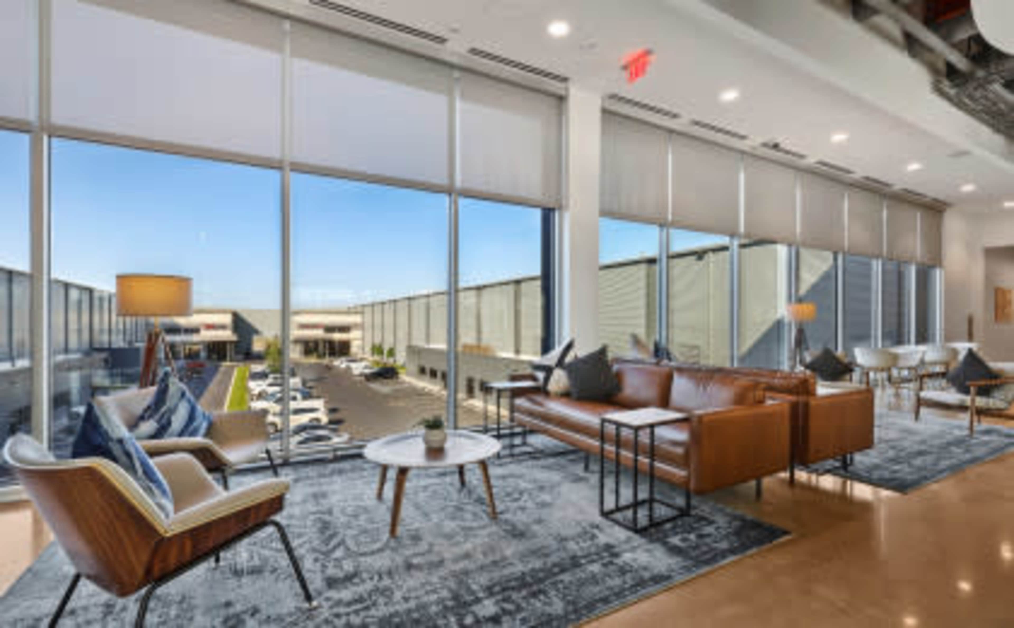 The image shows a modern lounge area with leather sofas, a rug, and large windows overlooking an outdoor parking lot.