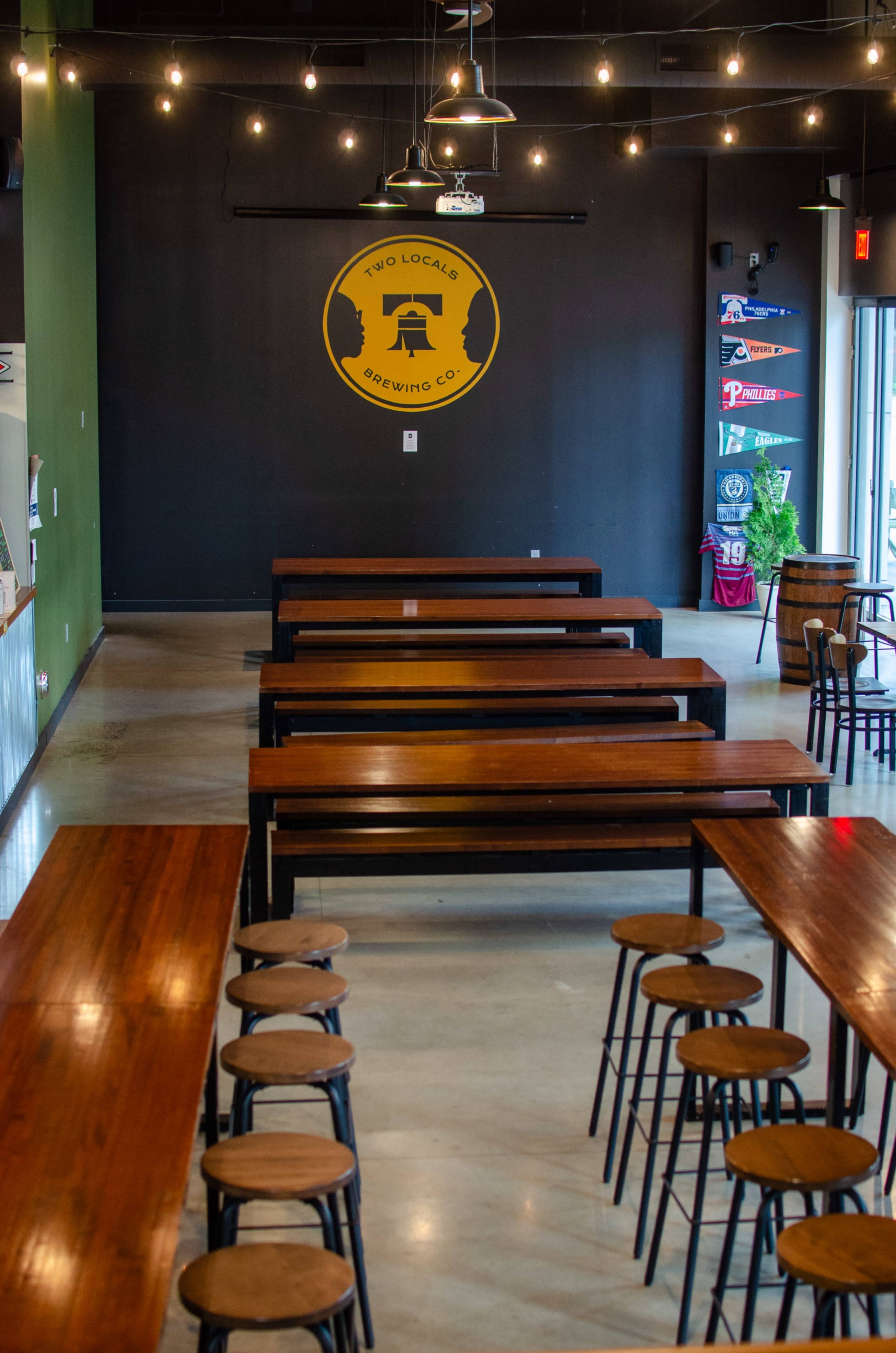 The image shows the interior of a brewing company with long wooden tables, high stools, and a large logo on the black wall.