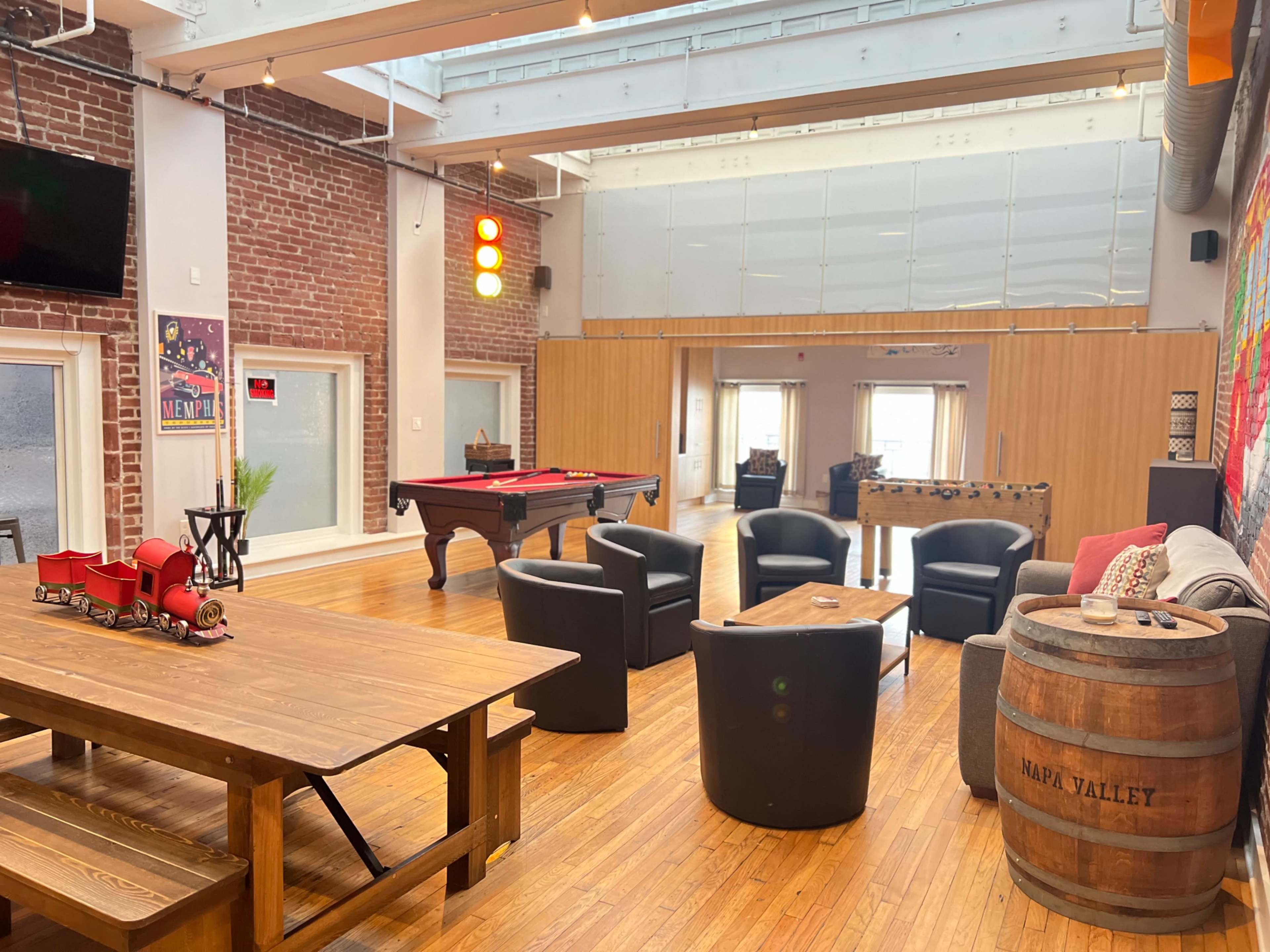 A spacious room with wooden flooring, featuring a pool table, foosball table, and seating areas with black chairs and a couch, alongside a large wooden dining table and a barrel.