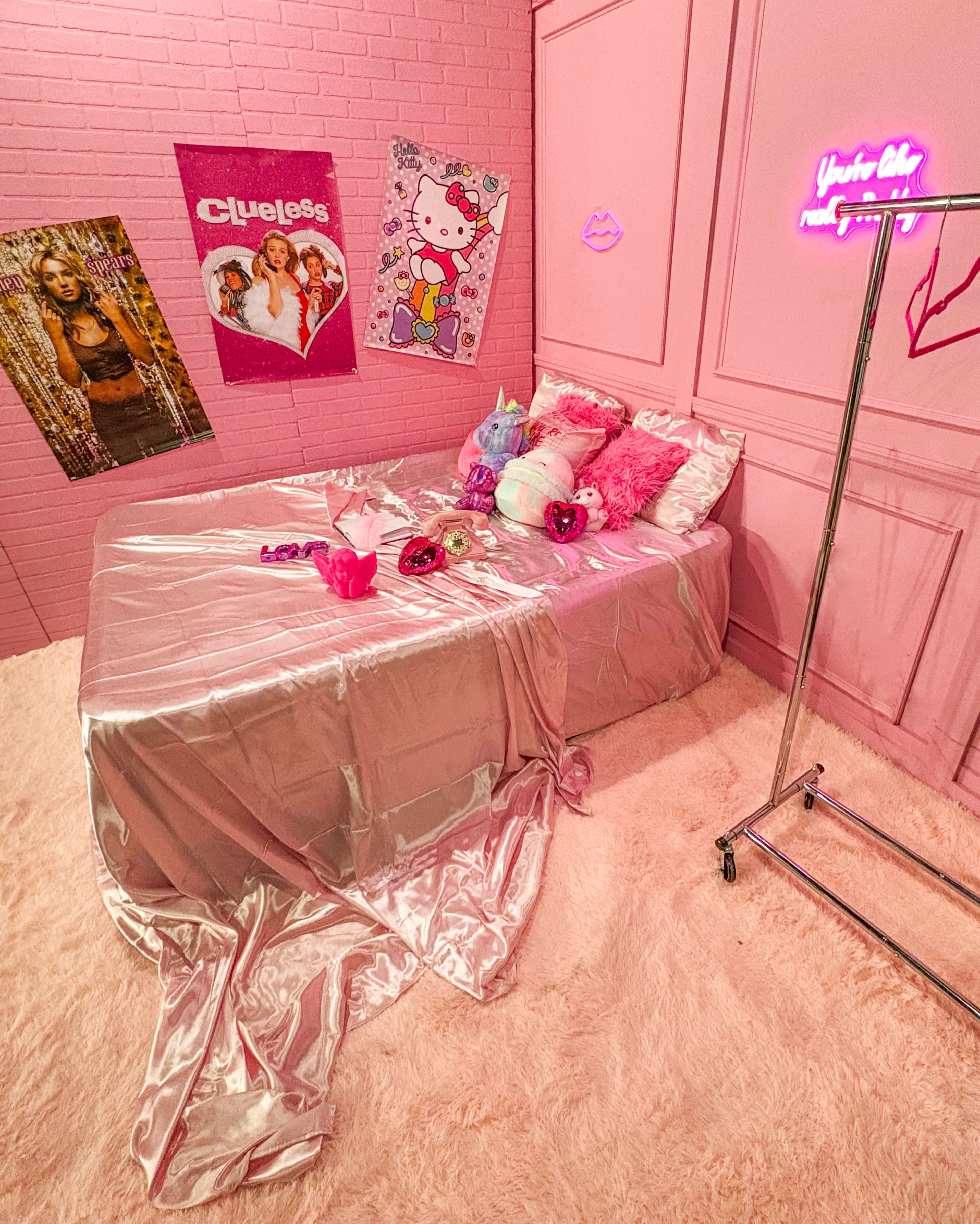 The image shows a pink-themed bedroom with a silk-covered bed, fluffy carpet, and decorated walls featuring posters and neon lights.