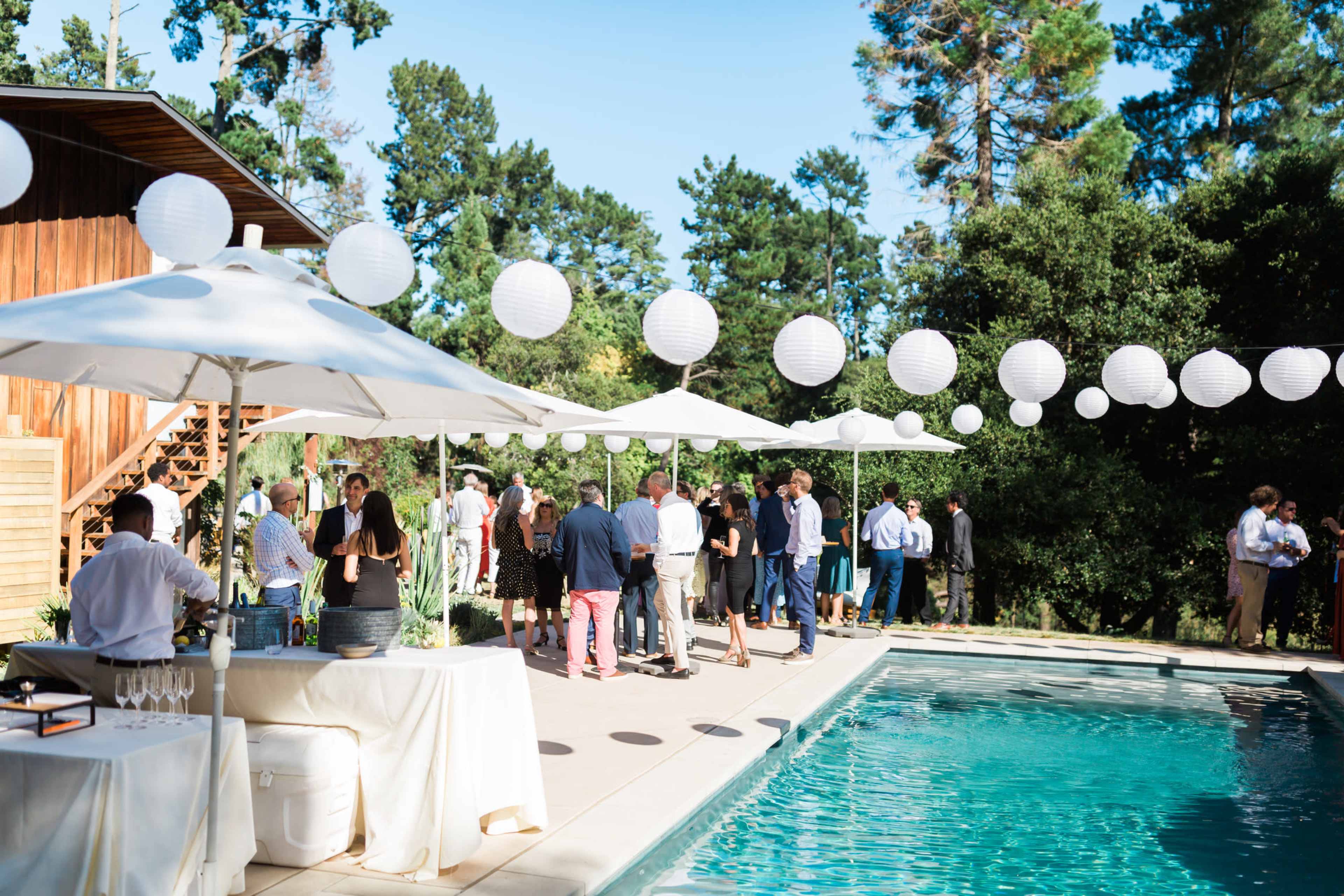 A gathering is taking place by a poolside with guests mingling under white lanterns and umbrellas, and a nearby building in the background.