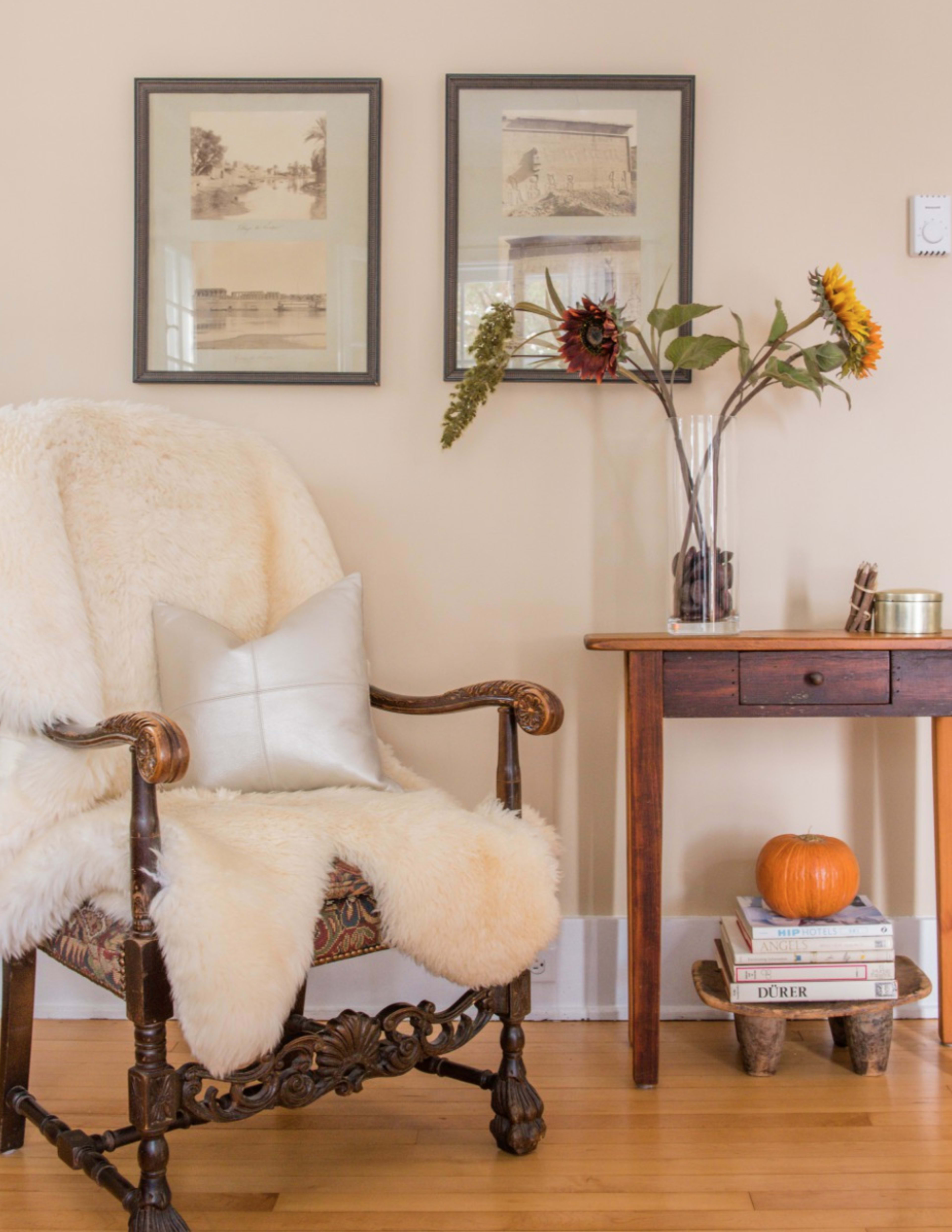 A wooden chair draped with a faux fur blanket sits beside a small table adorned with books, a pumpkin, and a vase of flowers against a beige wall featuring framed photographs.