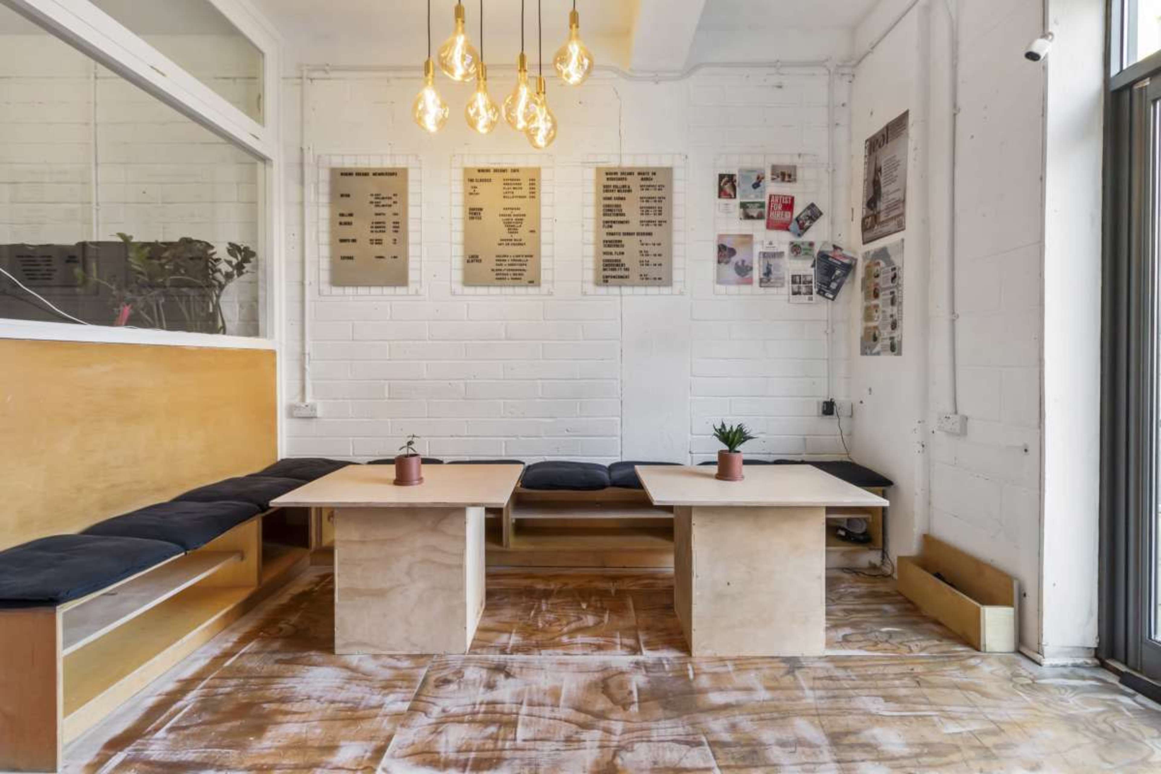 A minimalist café interior featuring wooden tables with potted plants, low seating against the wall, and pendant lights hanging from the ceiling.
