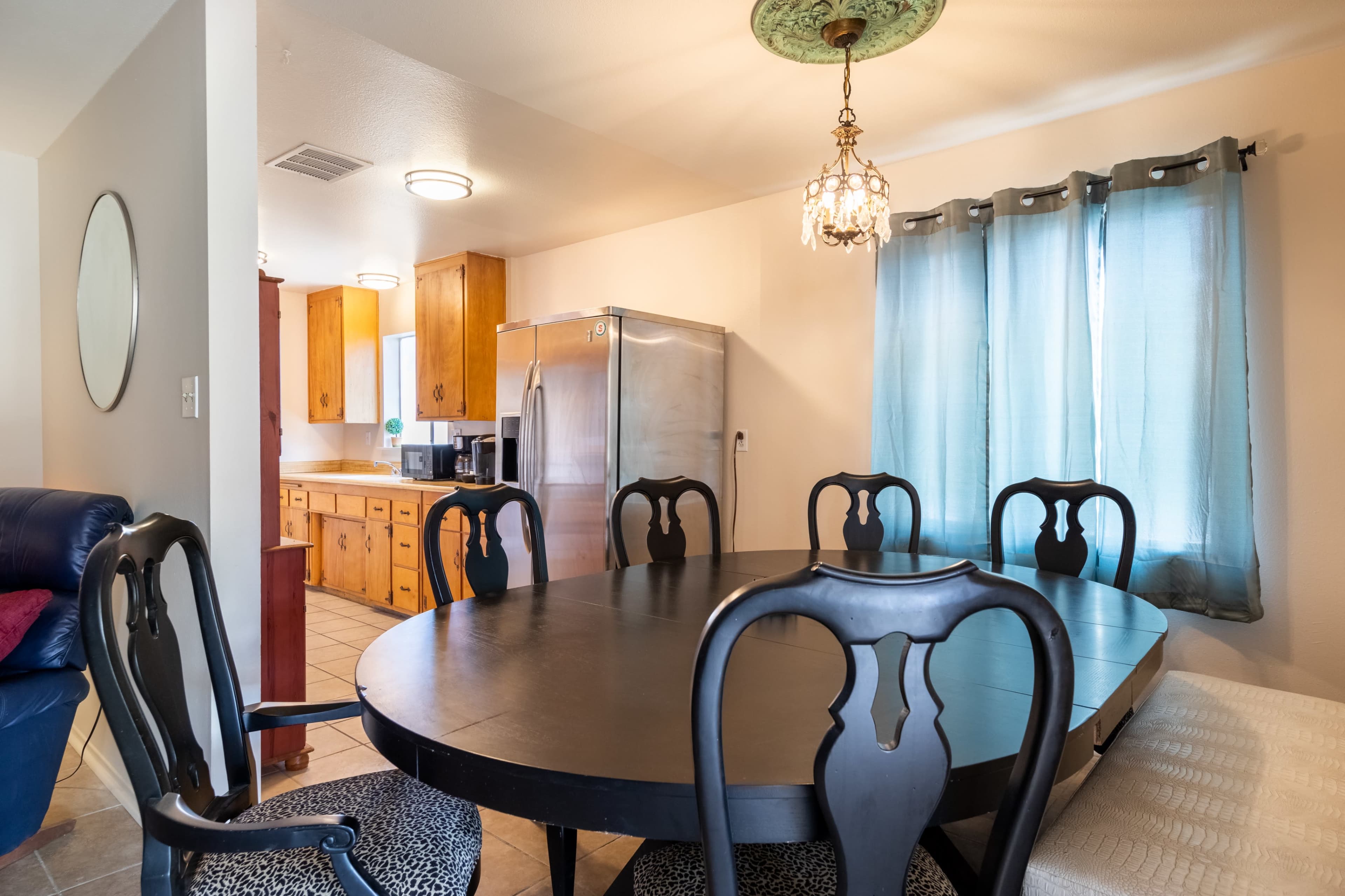 A round black dining table surrounded by chairs is positioned near a window with blue curtains in a kitchen area.