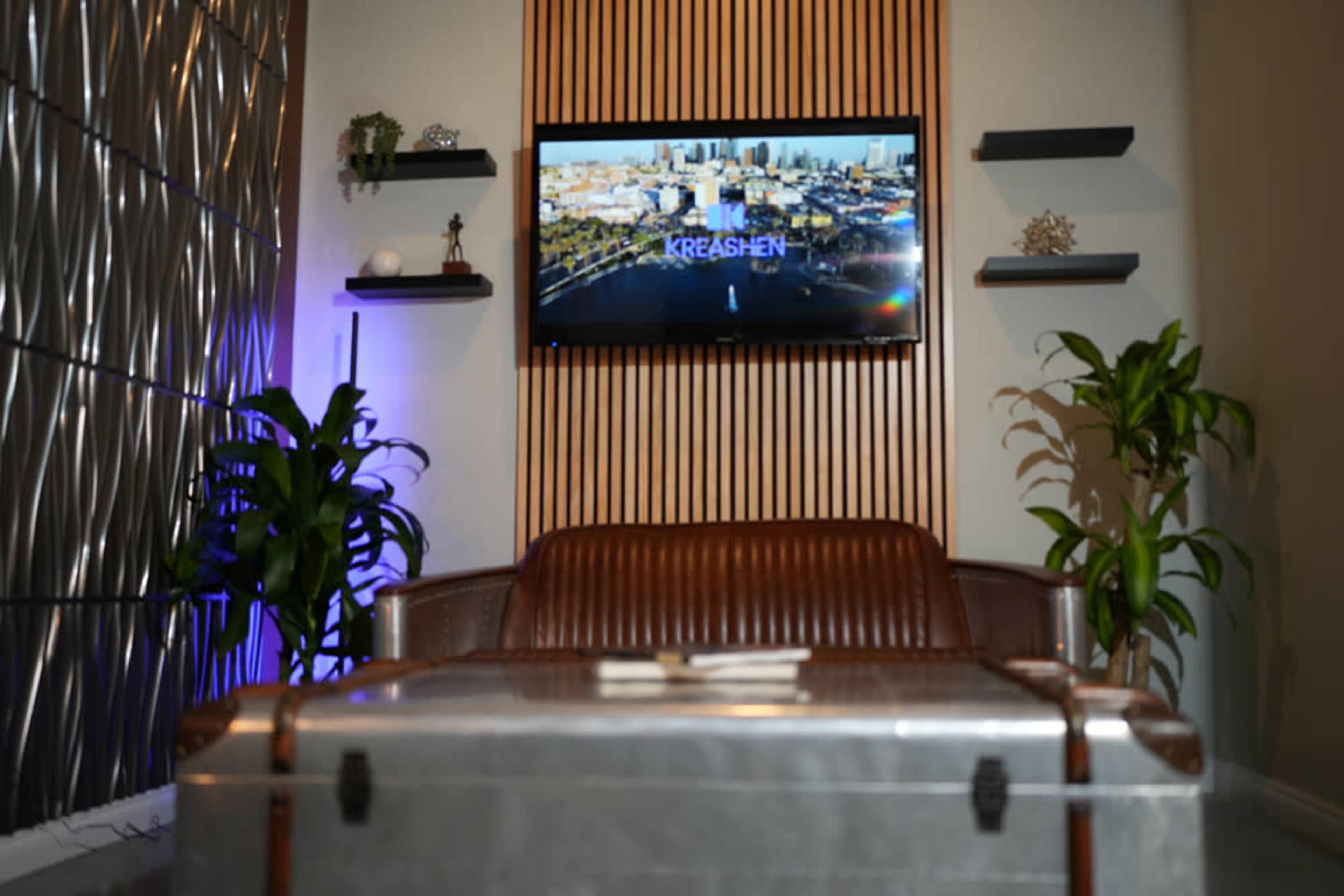 The image shows a modern interior space with a brown leather couch, an antique-style trunk as a coffee table, two green plants, and a television mounted on a wooden-paneled wall displaying a city landscape.