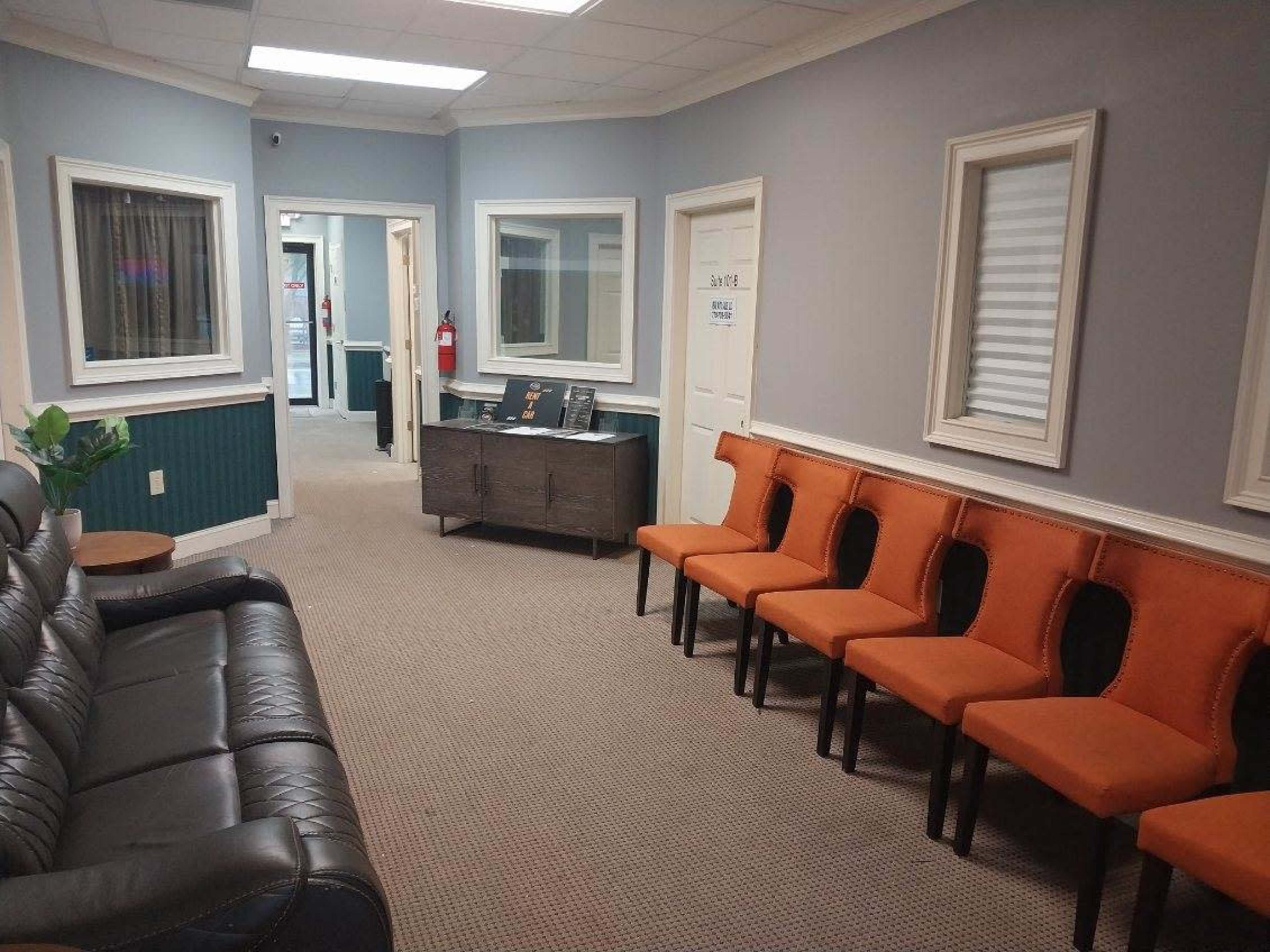 A waiting room with a black leather couch and a row of orange chairs against the wall, framed by light gray walls and two doors leading to other rooms.