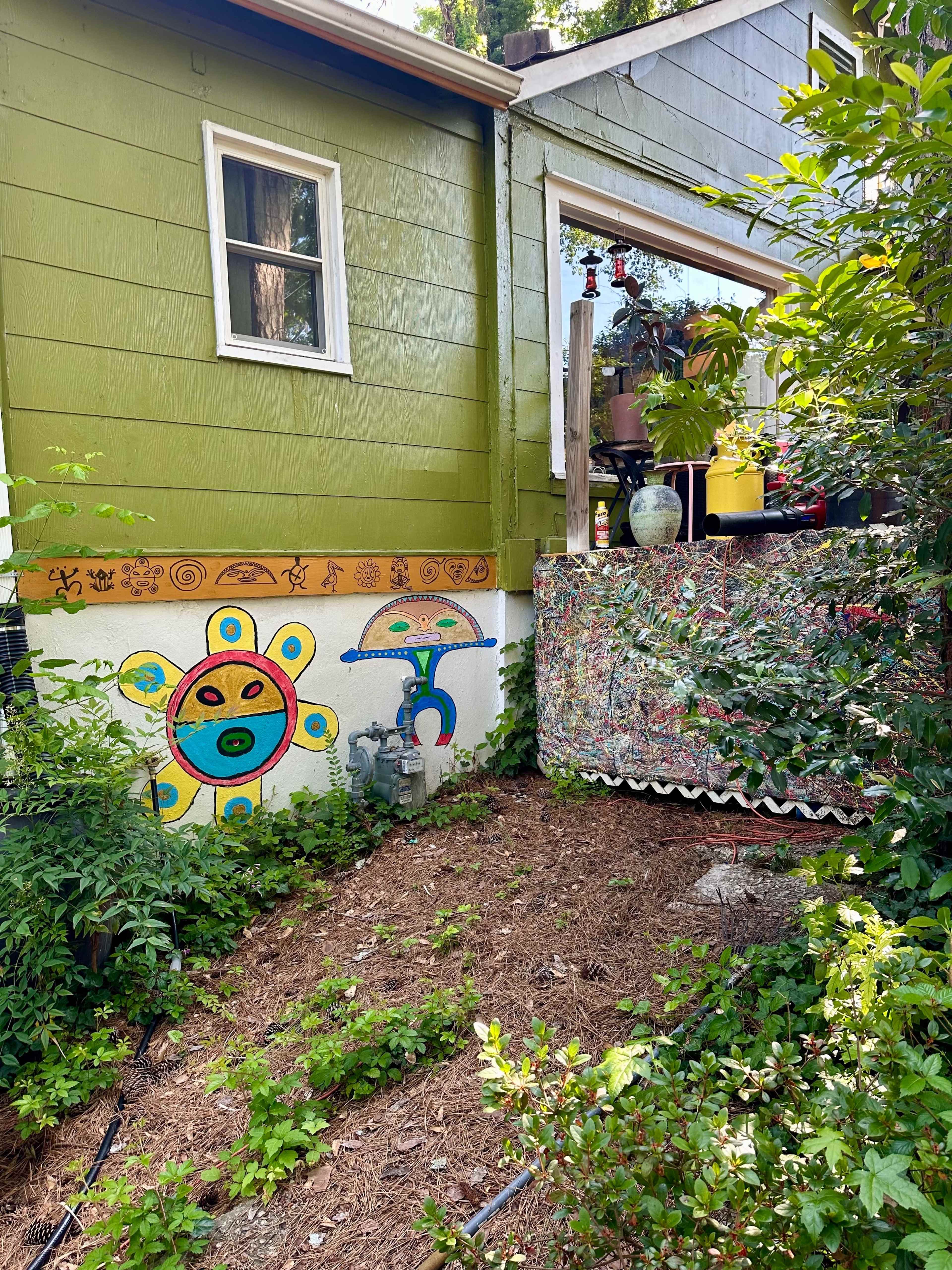A green house with a colorful mural on one side is surrounded by lush greenery and a decorative patio area.