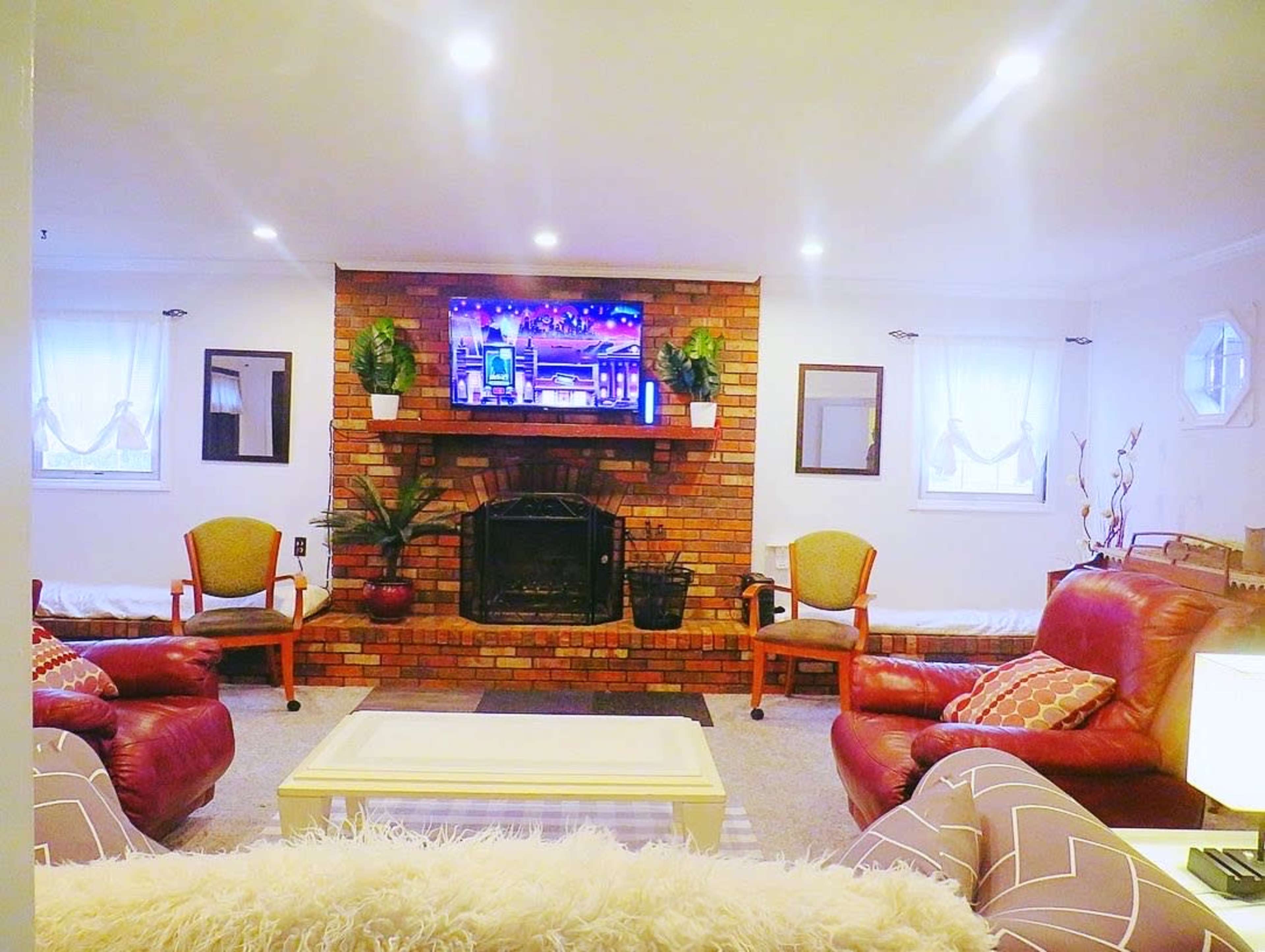 A cozy living room features a brick fireplace with a television above, flanked by two chairs and light-colored sofas.