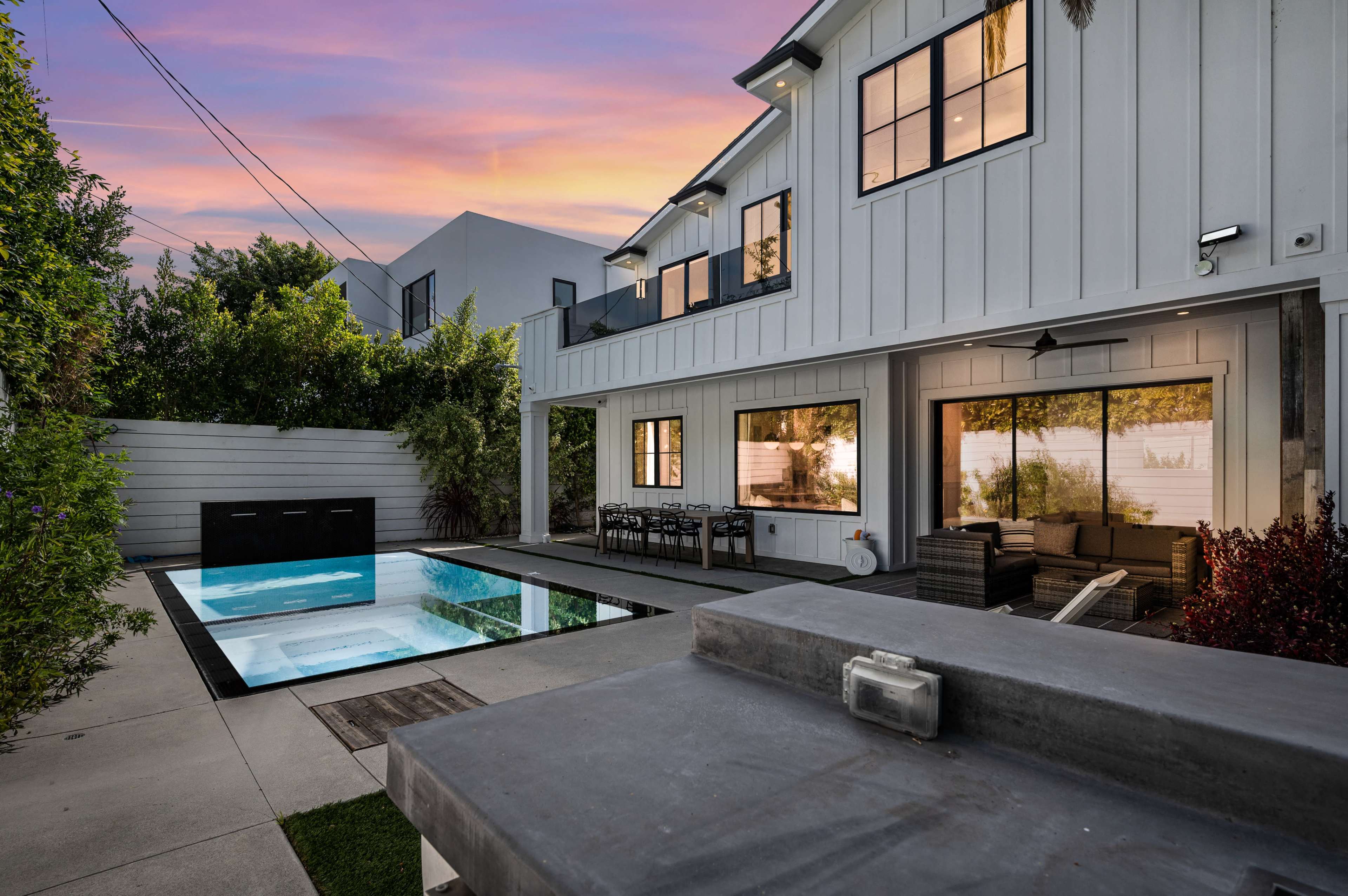 A modern backyard with a rectangular pool, a stone patio, and a two-story house featuring large windows.