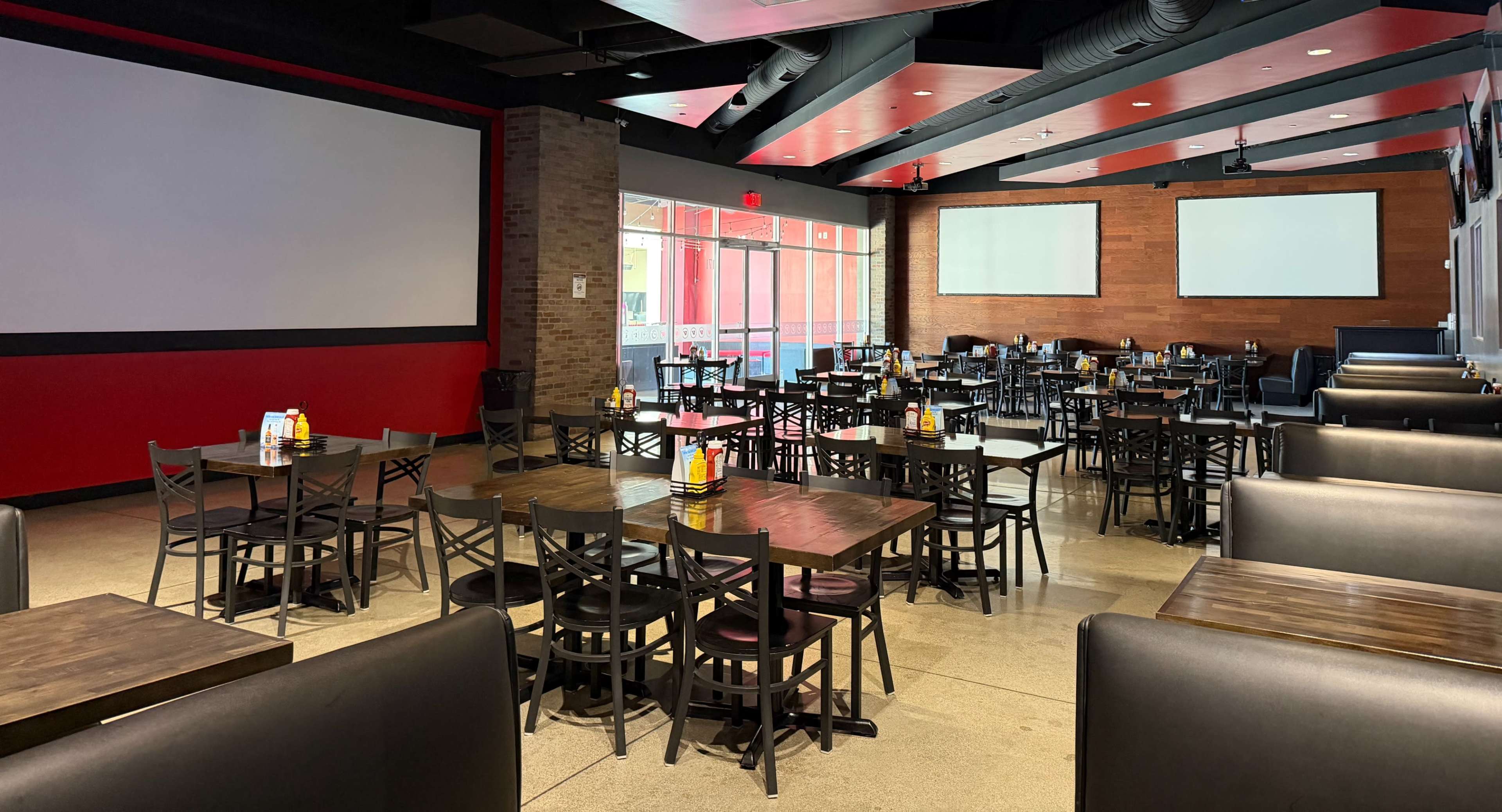 The image shows an empty restaurant interior with black chairs and wooden tables arranged neatly, featuring a mix of booth seating and open dining space, along with large screens on the walls.