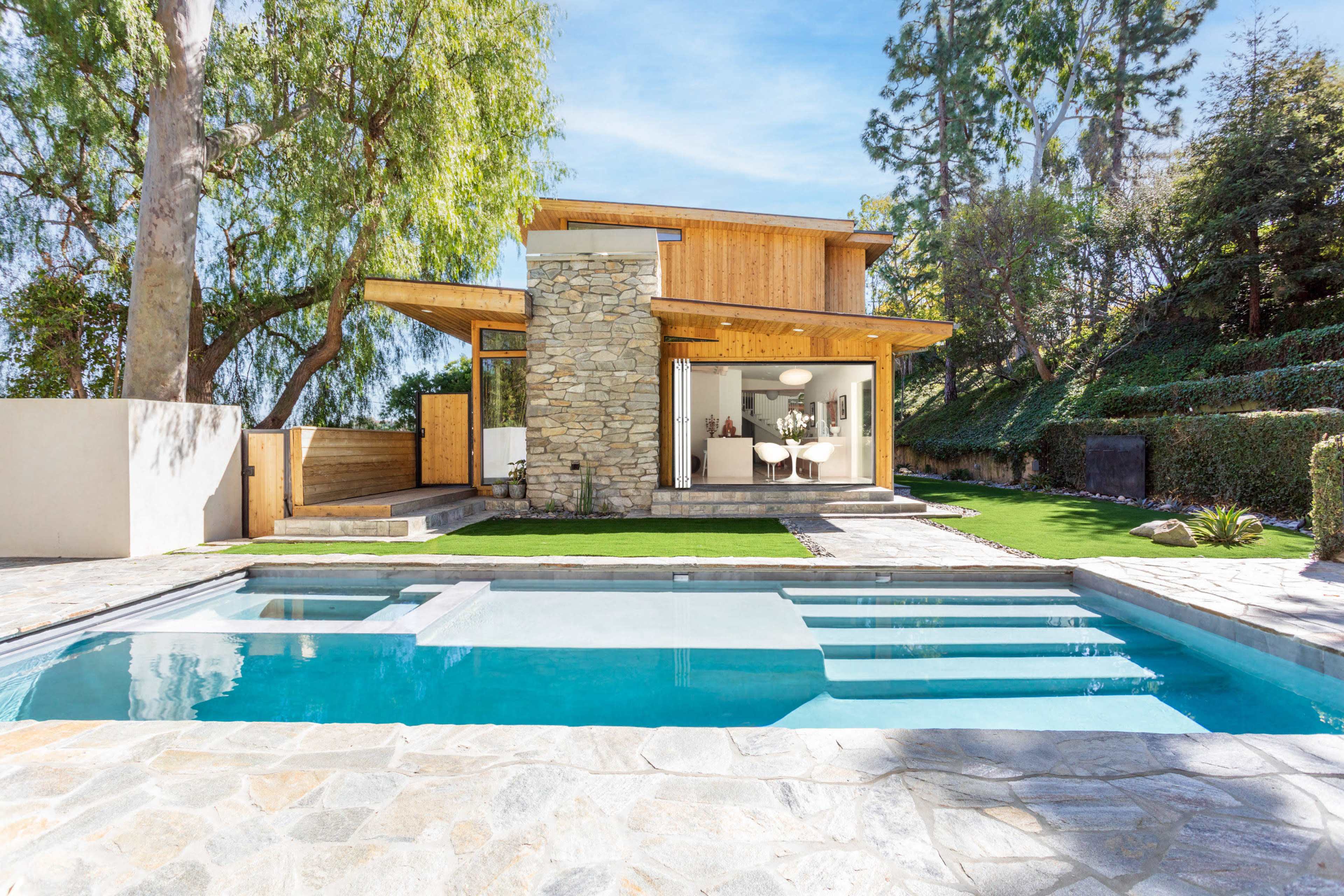 A modern house with a stone facade features large windows and a pool in the foreground, surrounded by landscaped greenery.