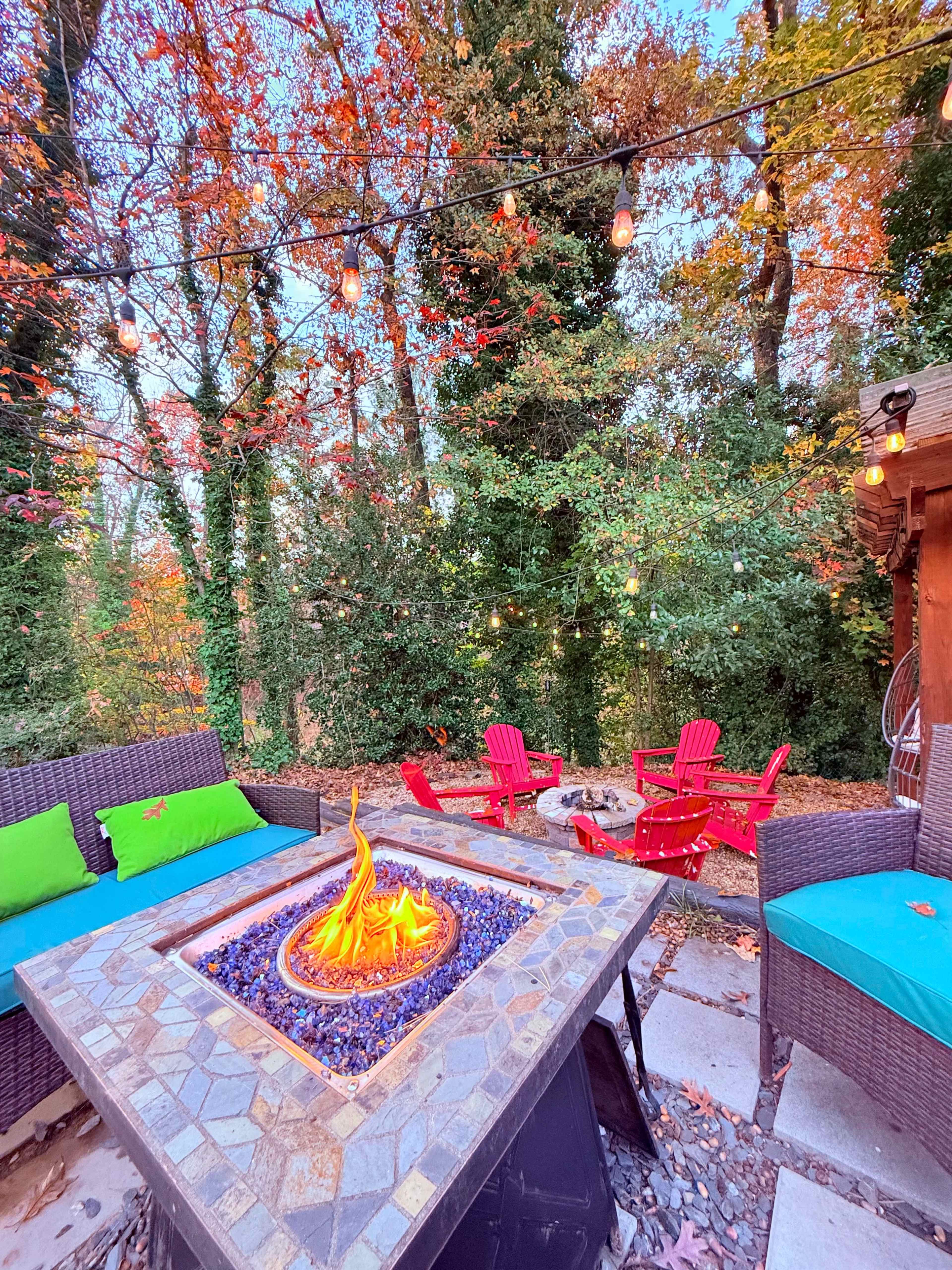 A fire pit with flames sits at the center of a cozy outdoor seating area surrounded by colorful fall foliage and illuminated by string lights.
