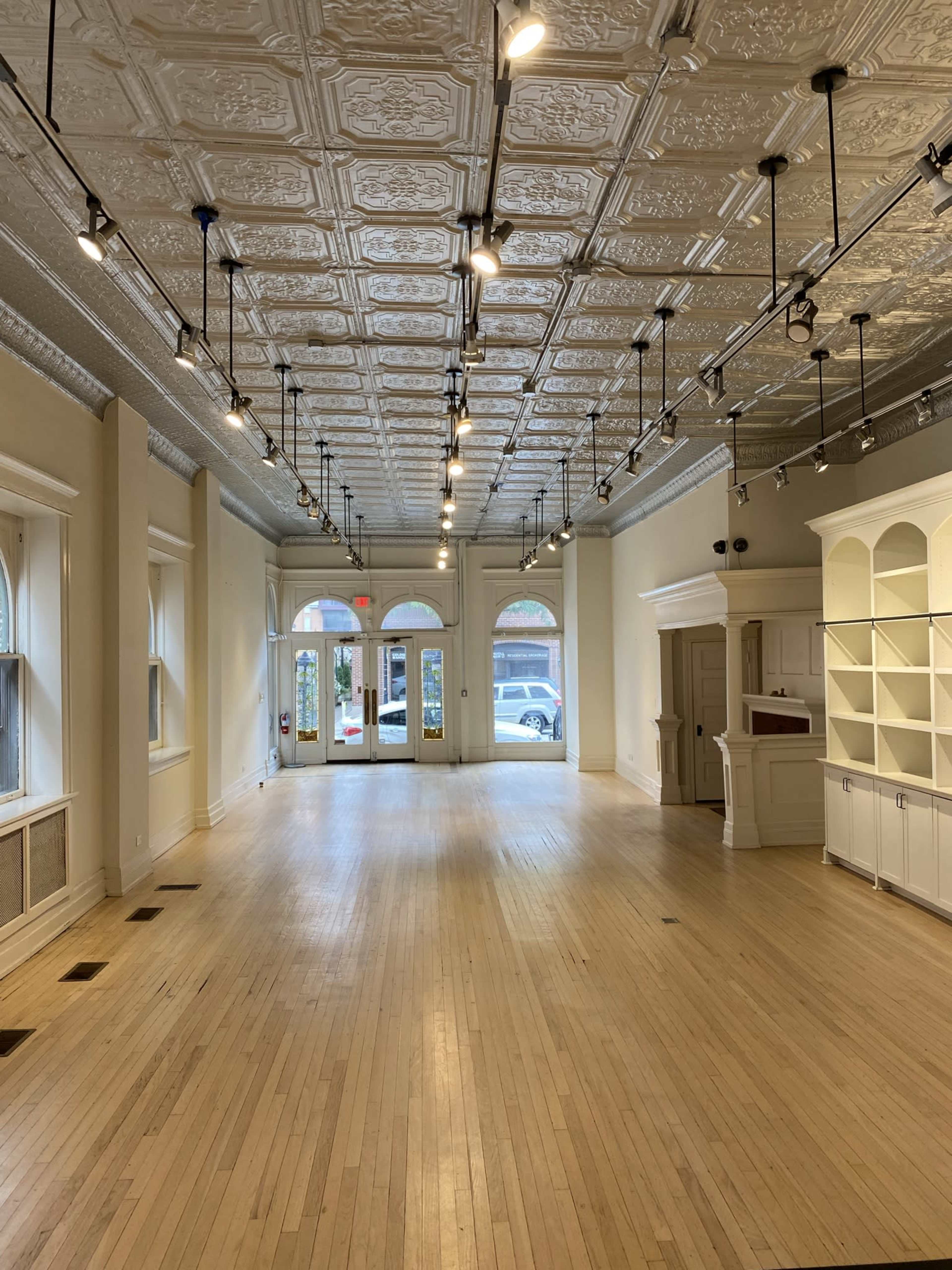 An empty retail space with a wooden floor, decorative ceiling tiles, and large front windows.