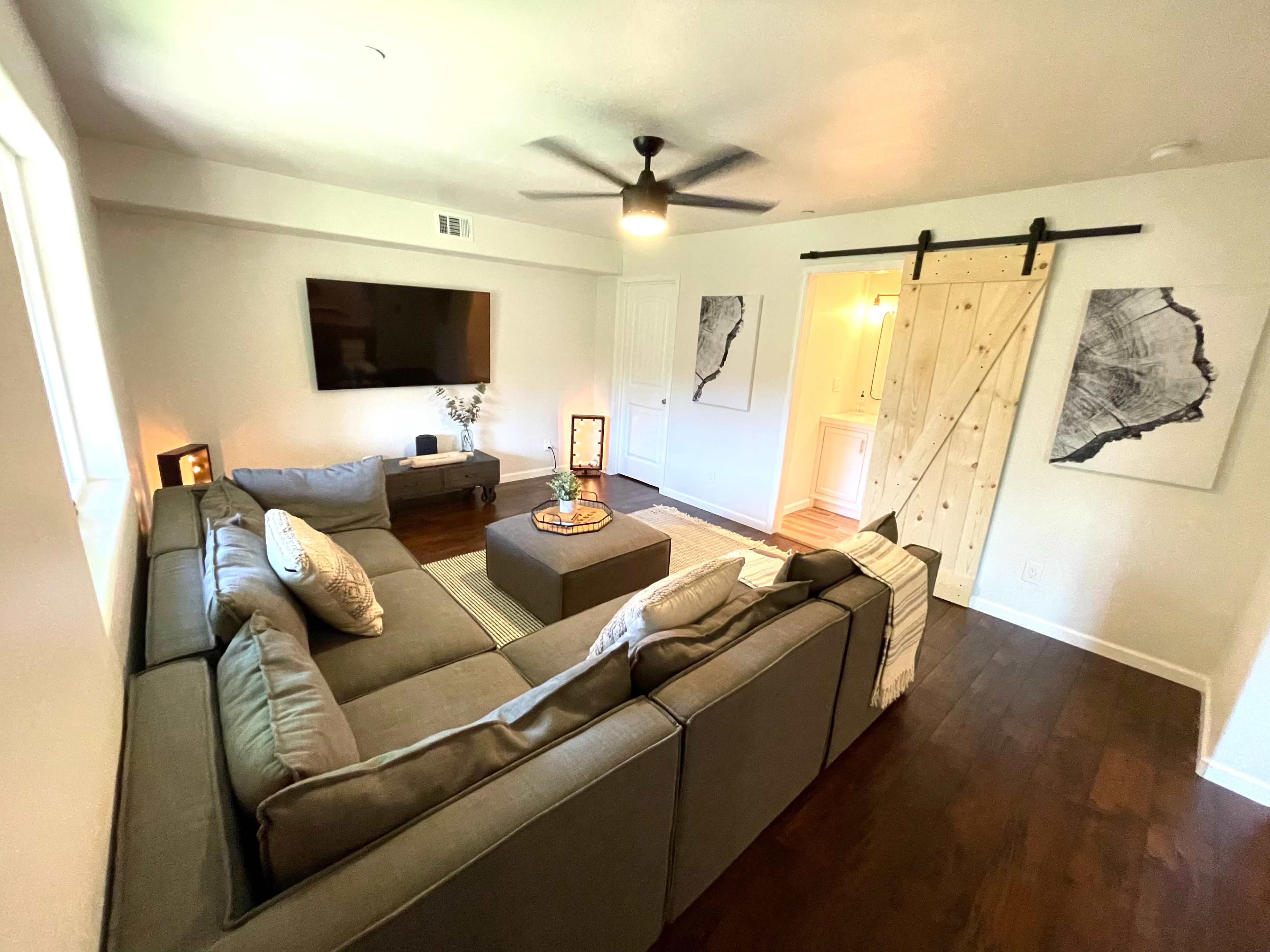 A modern living room features a gray sectional couch, a central coffee table, a wall-mounted television, and a sliding barn door leading to another room.
