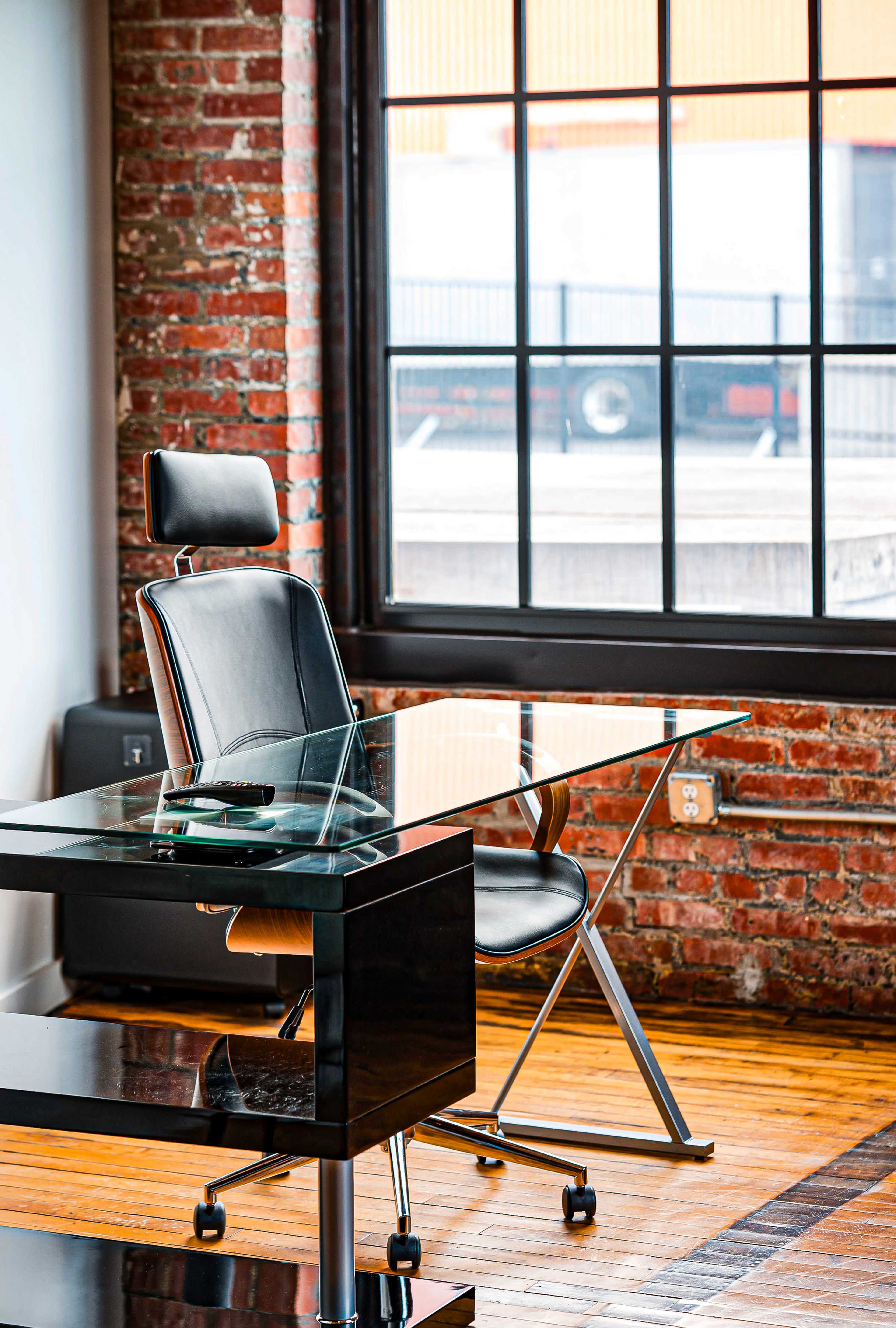 A modern black desk with a glass top and an ergonomic black chair in a room with exposed brick walls and large windows.