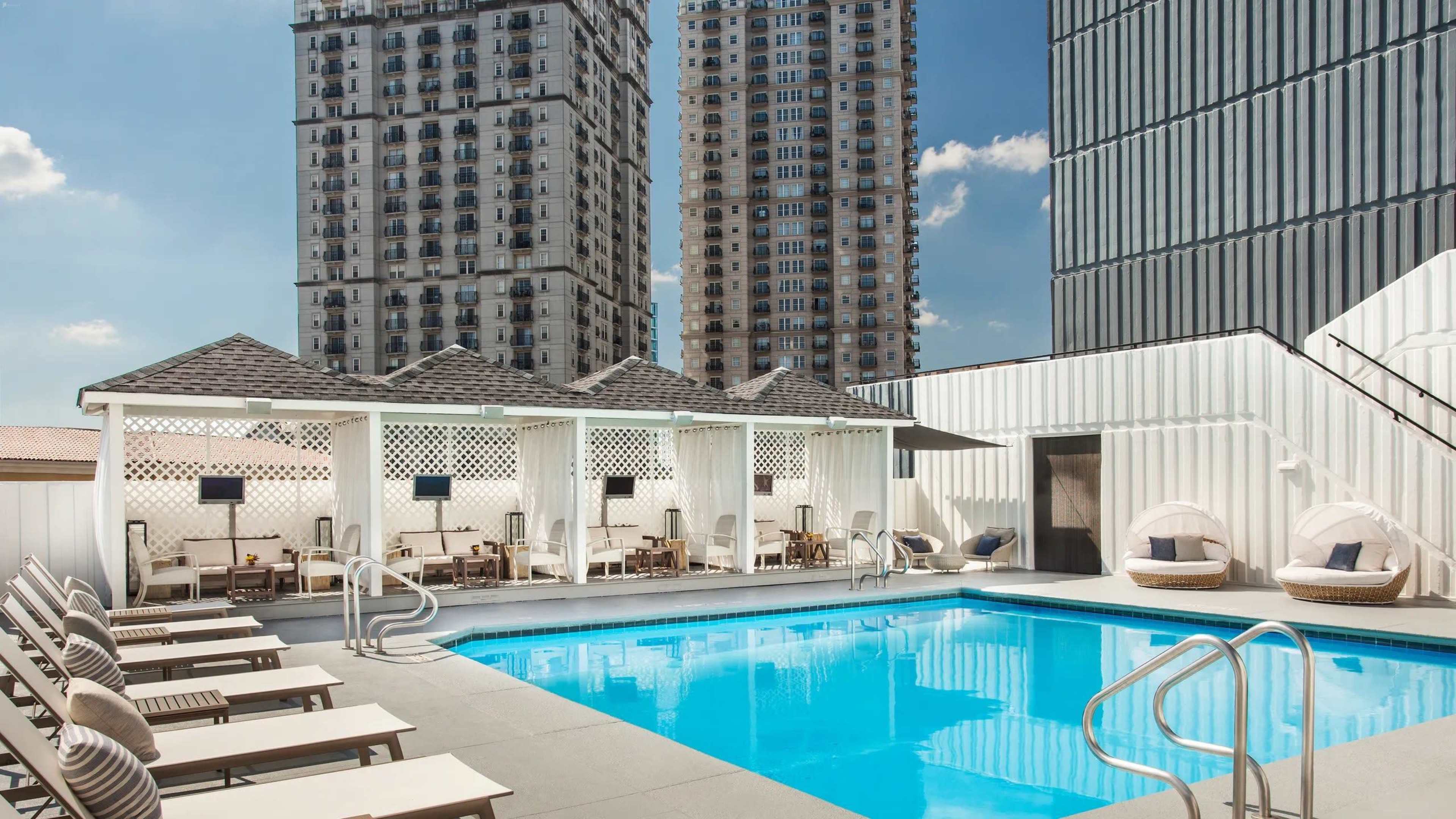 The image shows a modern rooftop pool area surrounded by tall apartment buildings, with lounge chairs and cabanas.