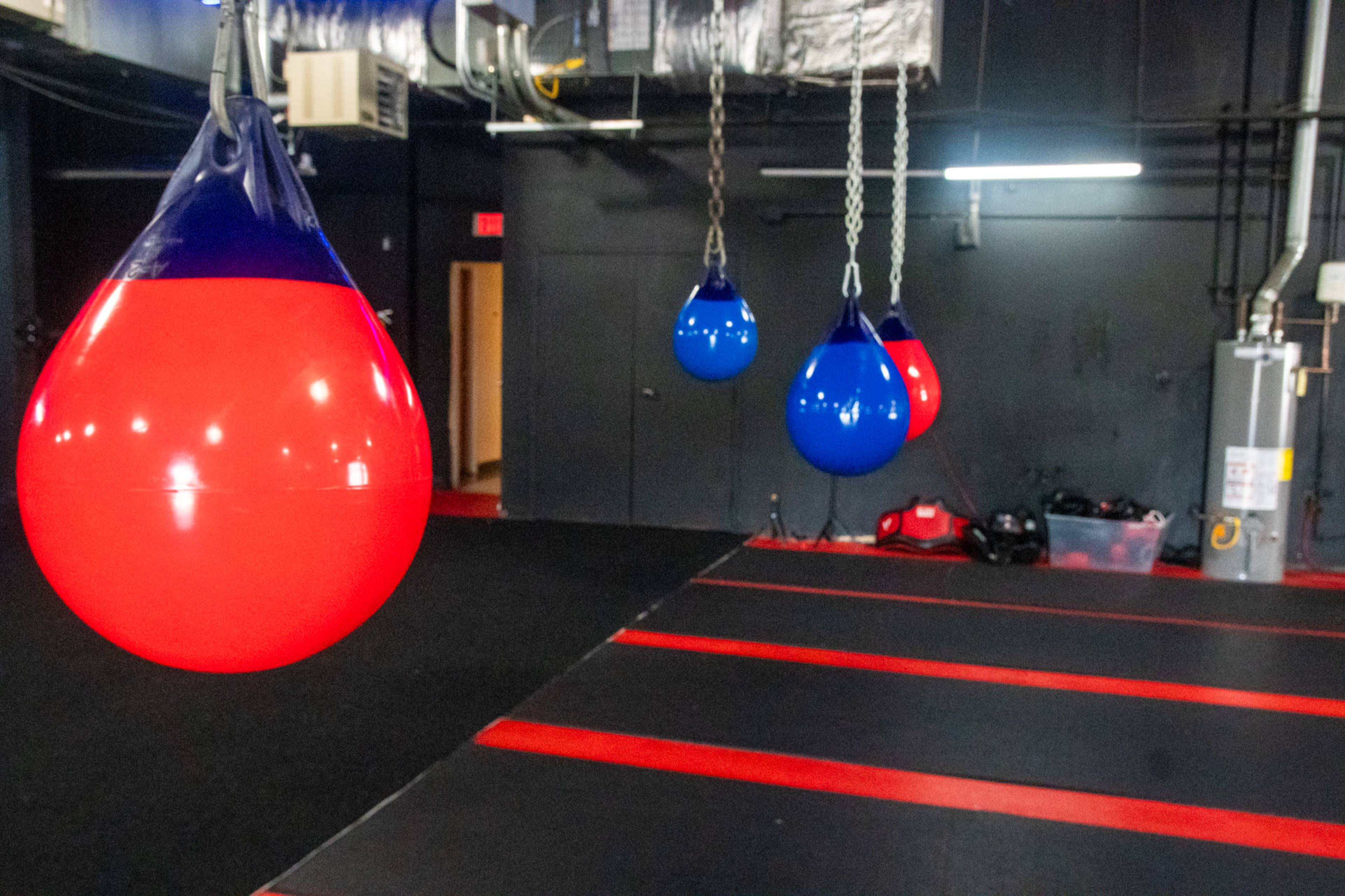 The image shows a training area with red and blue punching bags hanging from the ceiling, set against a black floor with red stripes.
