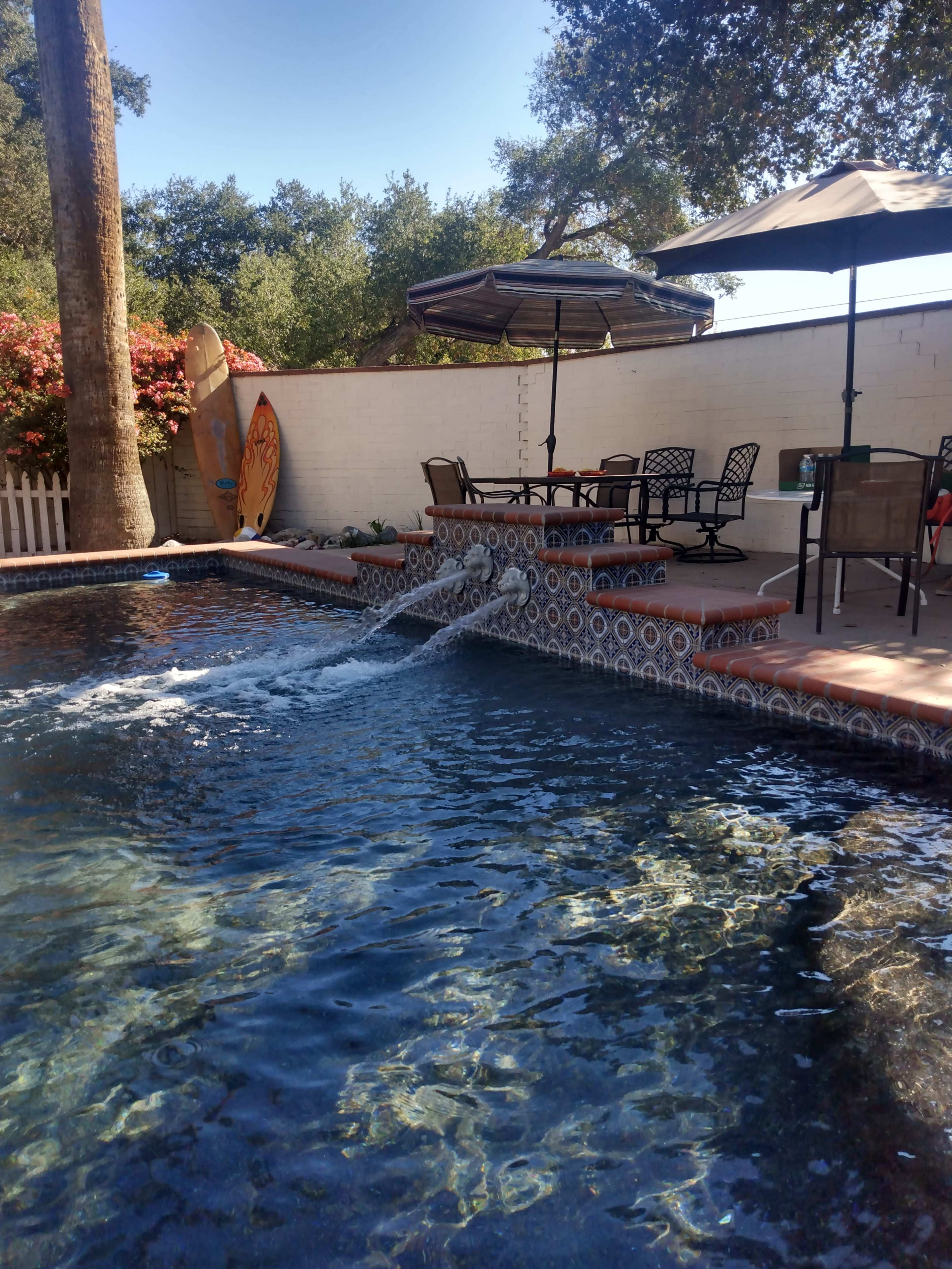 A swimming pool with a fountain feature is surrounded by patio furniture and shaded by umbrellas, with a surfboard resting against a wall in the background.