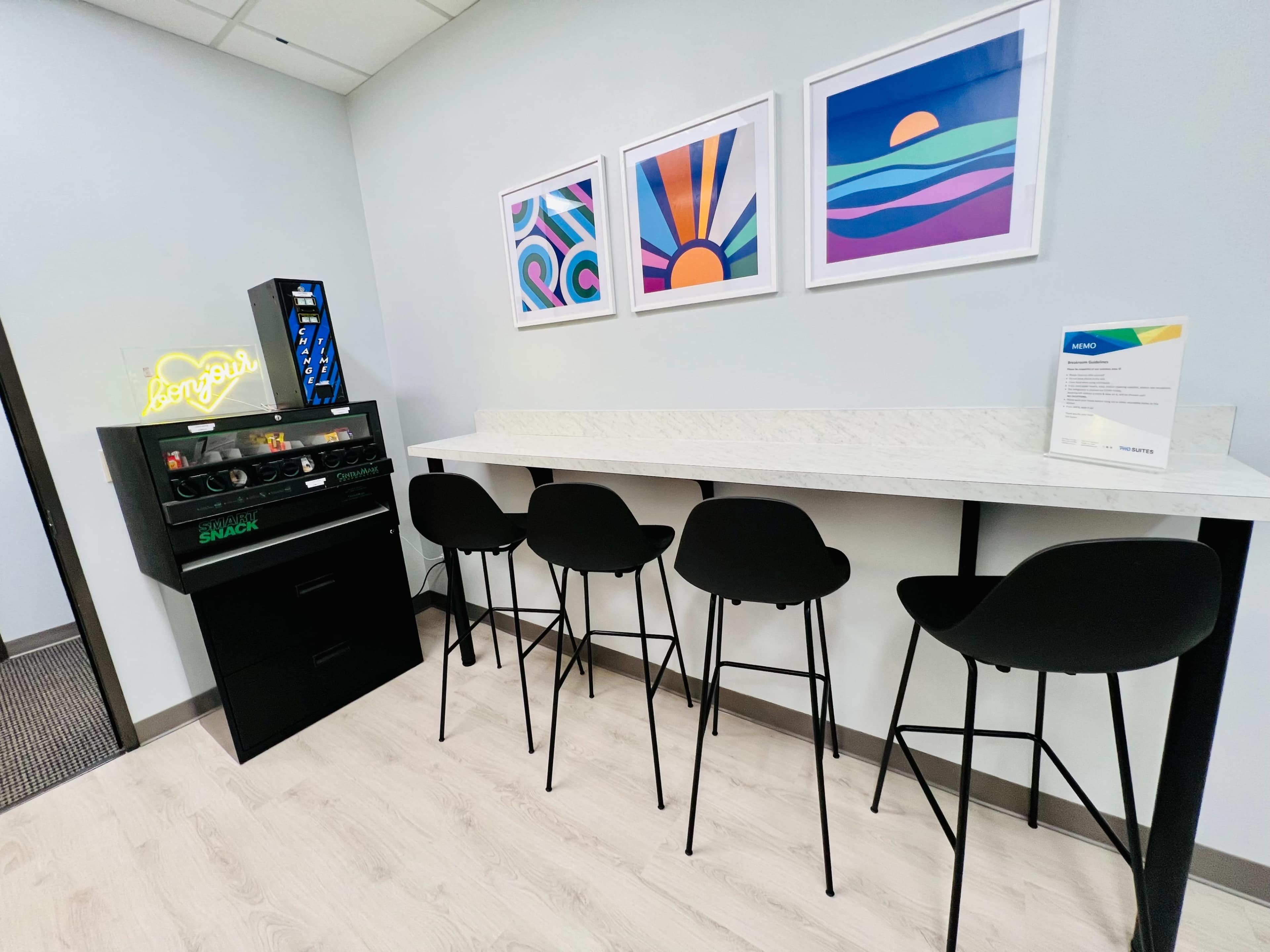 A modern break area features a countertop with four black chairs, a snack vending machine, and colorful framed art on the wall.
