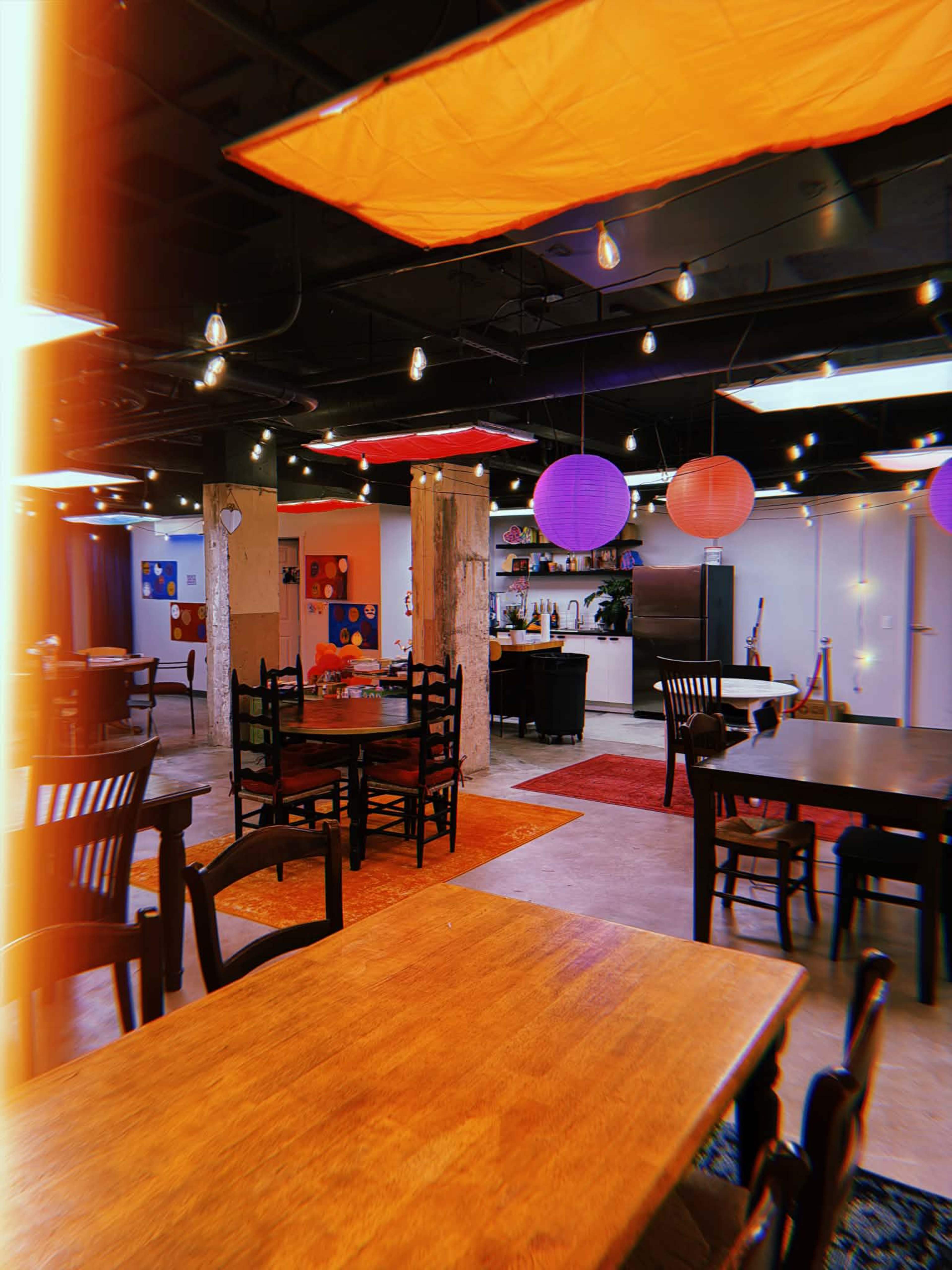 A spacious dining area features wooden tables, colorful hanging lanterns, and exposed brick walls, with various seating arrangements under soft lighting.
