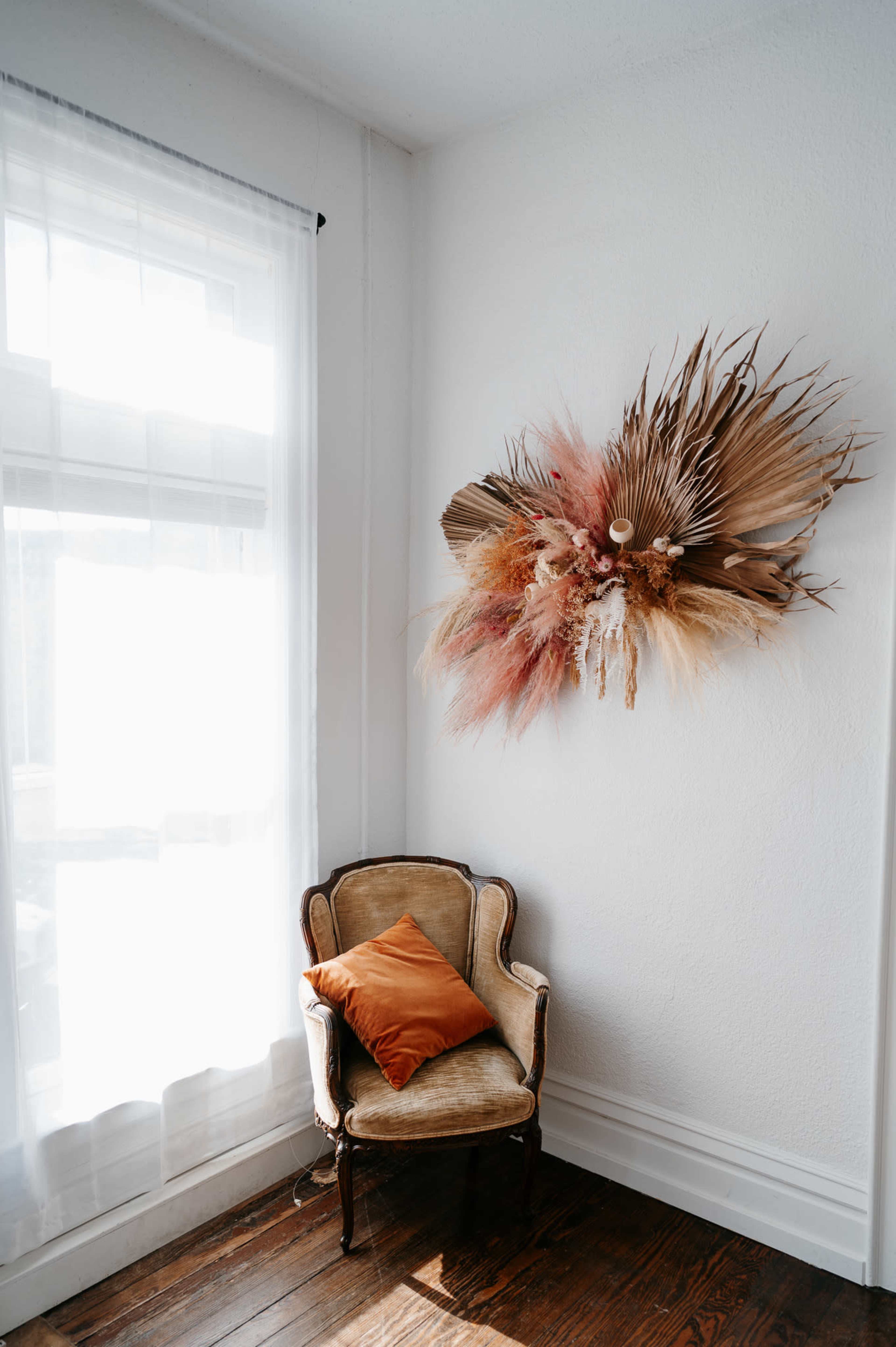 A vintage chair with an orange pillow sits beside a window, while a large wall arrangement of dried plants decorates the white wall.