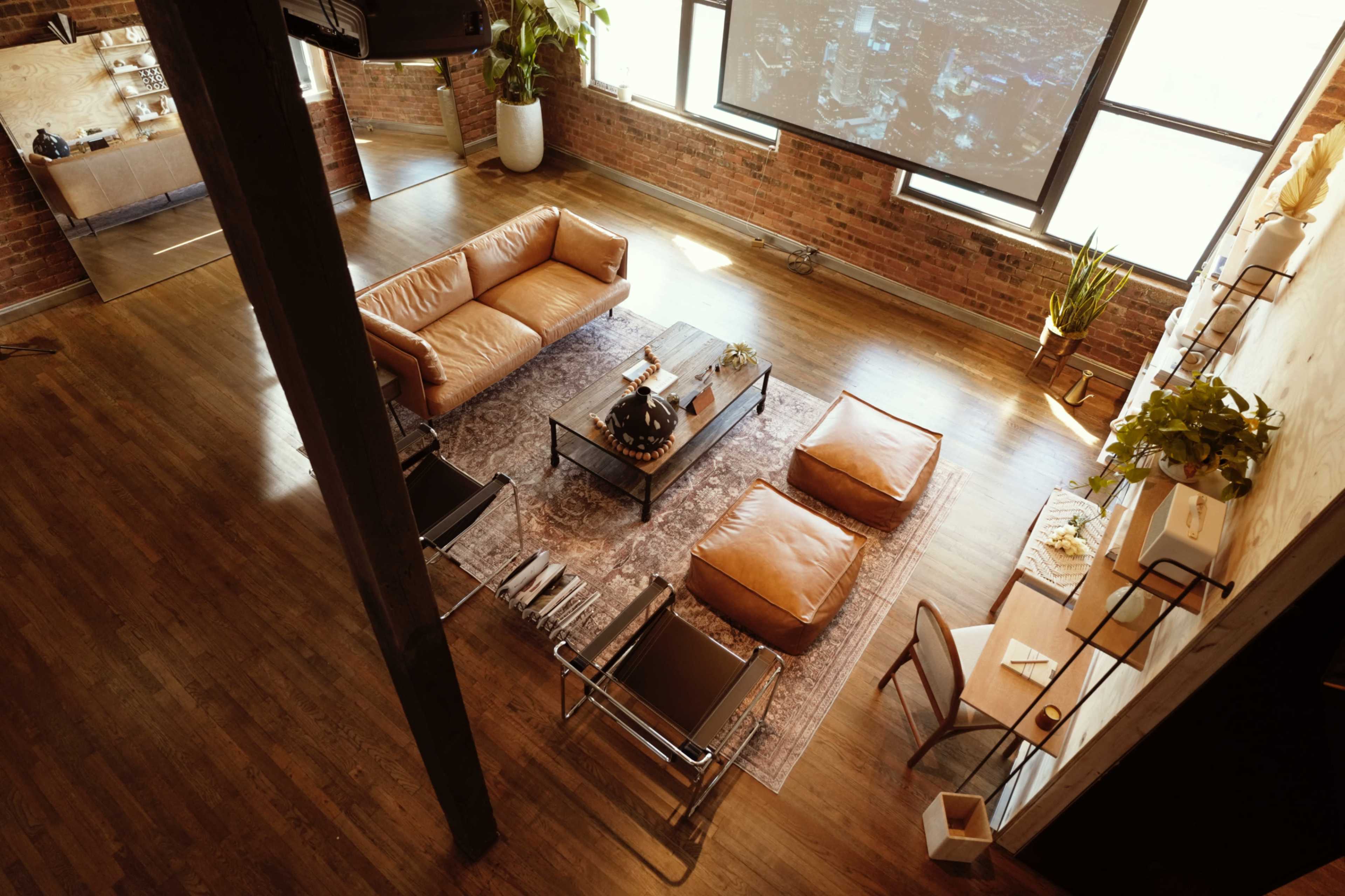 The image shows a spacious living room with a leather sofa, a coffee table, and two chairs, set against a backdrop of exposed brick walls and large windows.