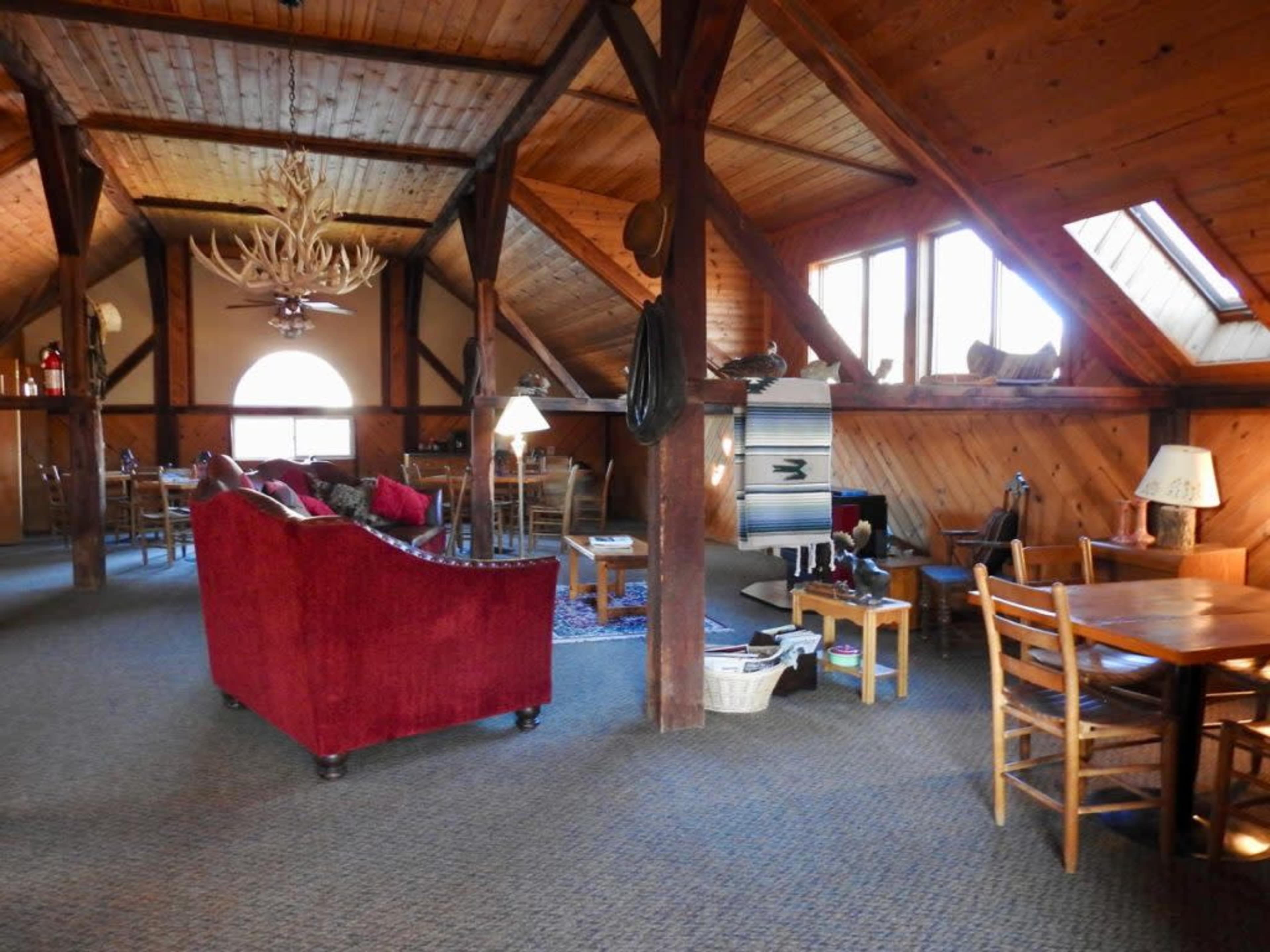 A spacious wooden interior with high ceilings, featuring a red sofa, various wooden furniture, and large windows allowing natural light to enter.