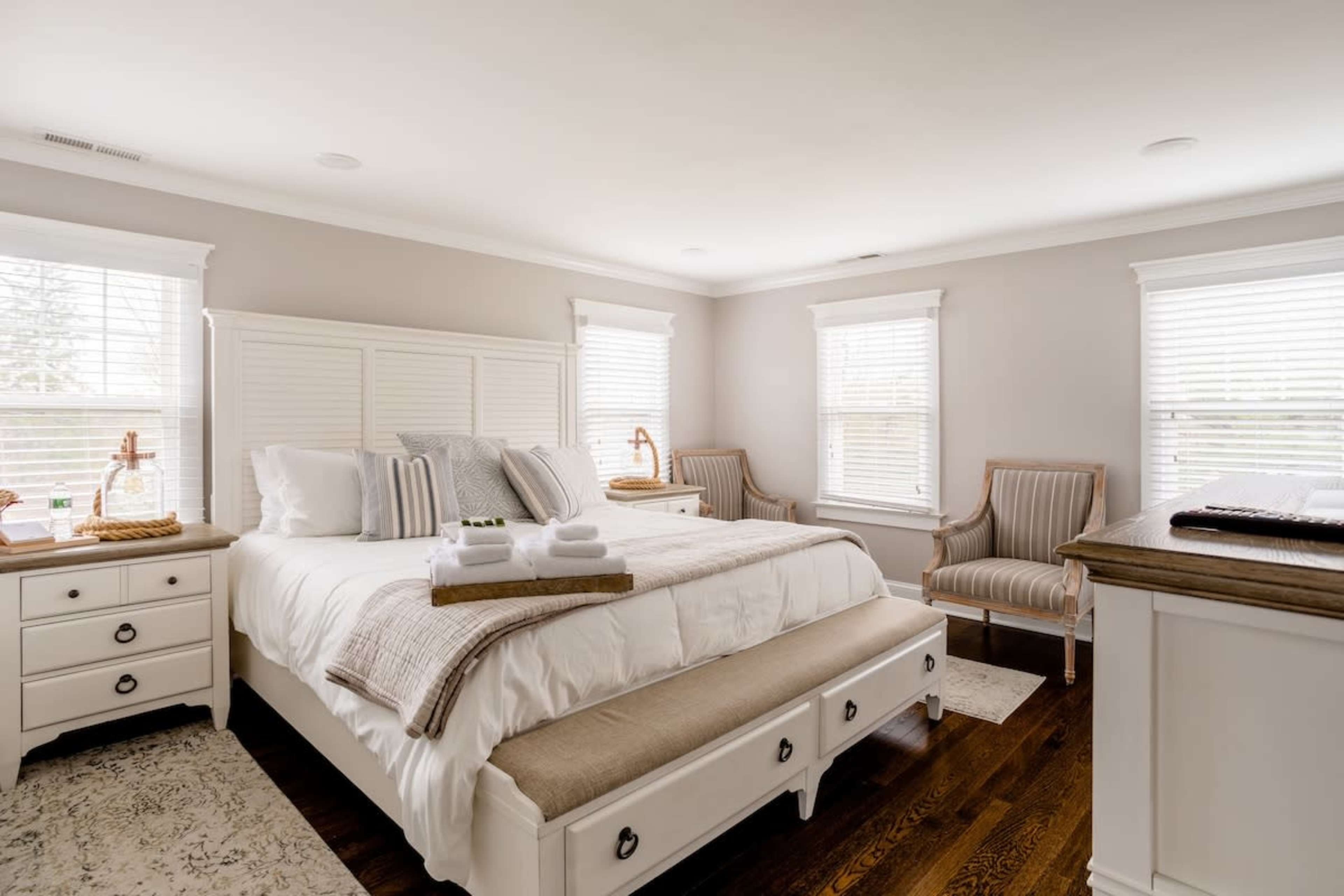 A furnished bedroom with a bed, two nightstands, seating chairs, and large windows allowing natural light.