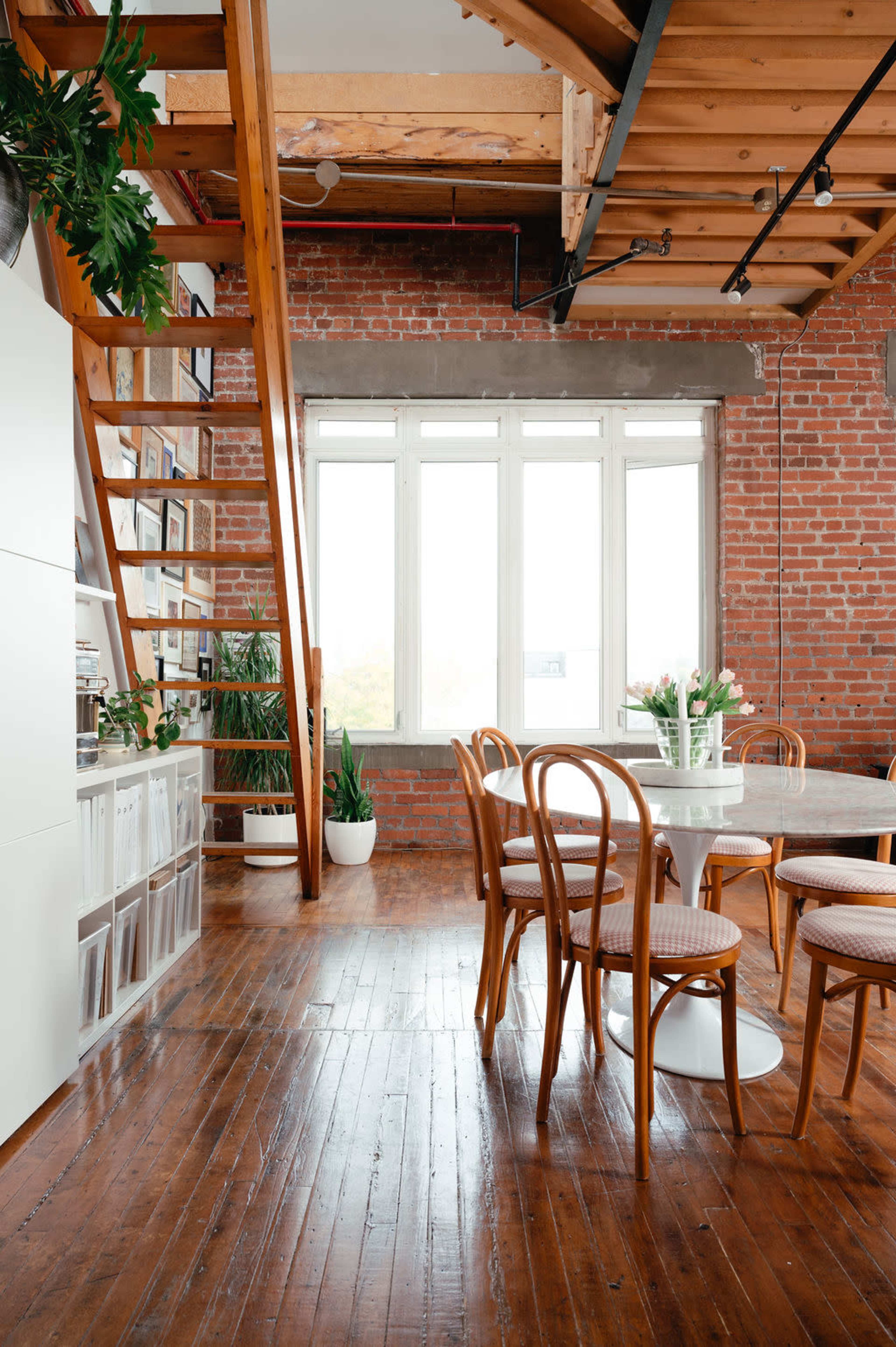 A bright, airy dining area with a round table surrounded by bentwood chairs, a staircase leading to a loft, and exposed brick walls.