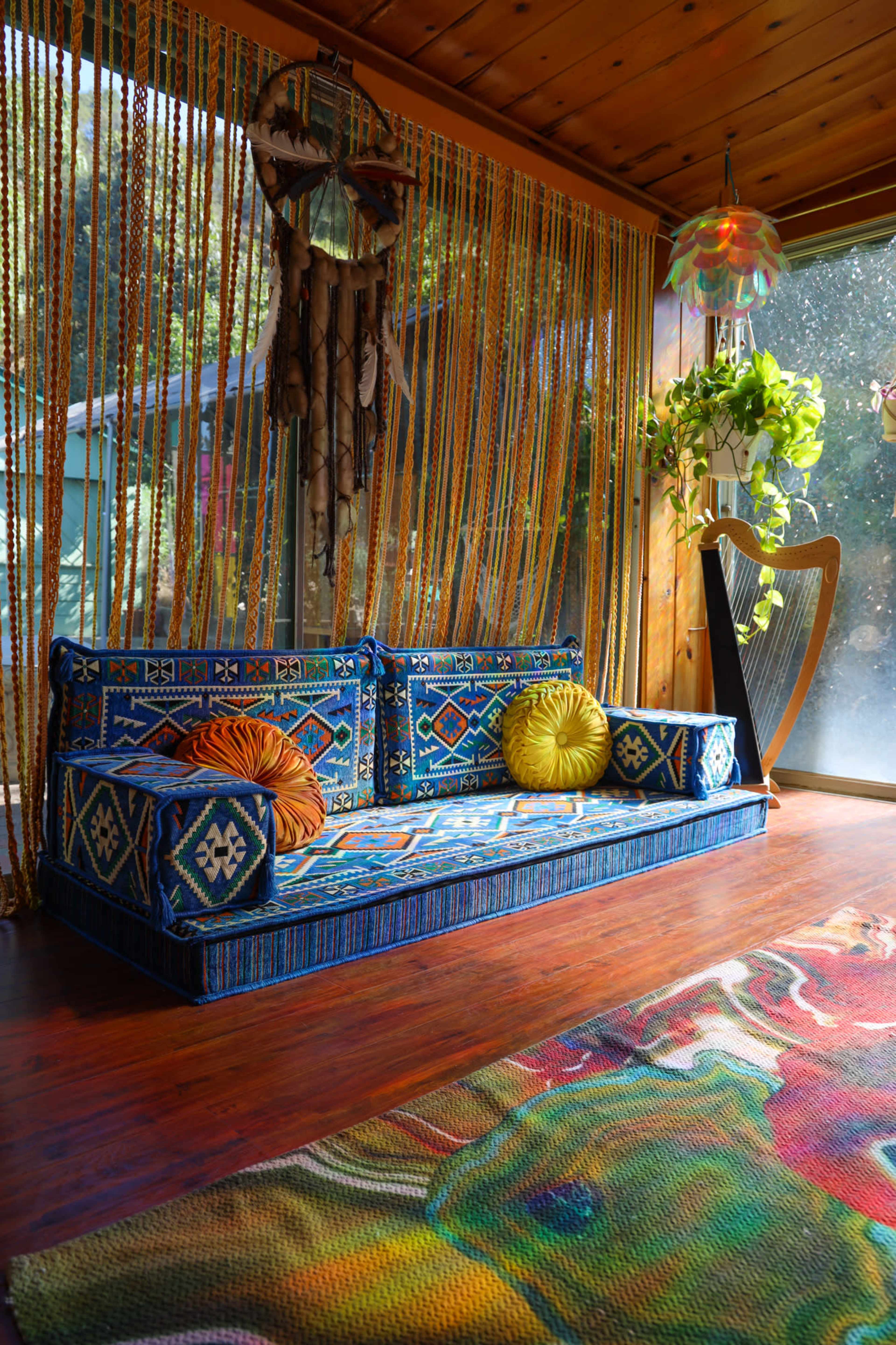 A colorful, patterned couch with decorative pillows in a room adorned with textured wall hangings and a large plant, alongside a harp and a vibrant rug.