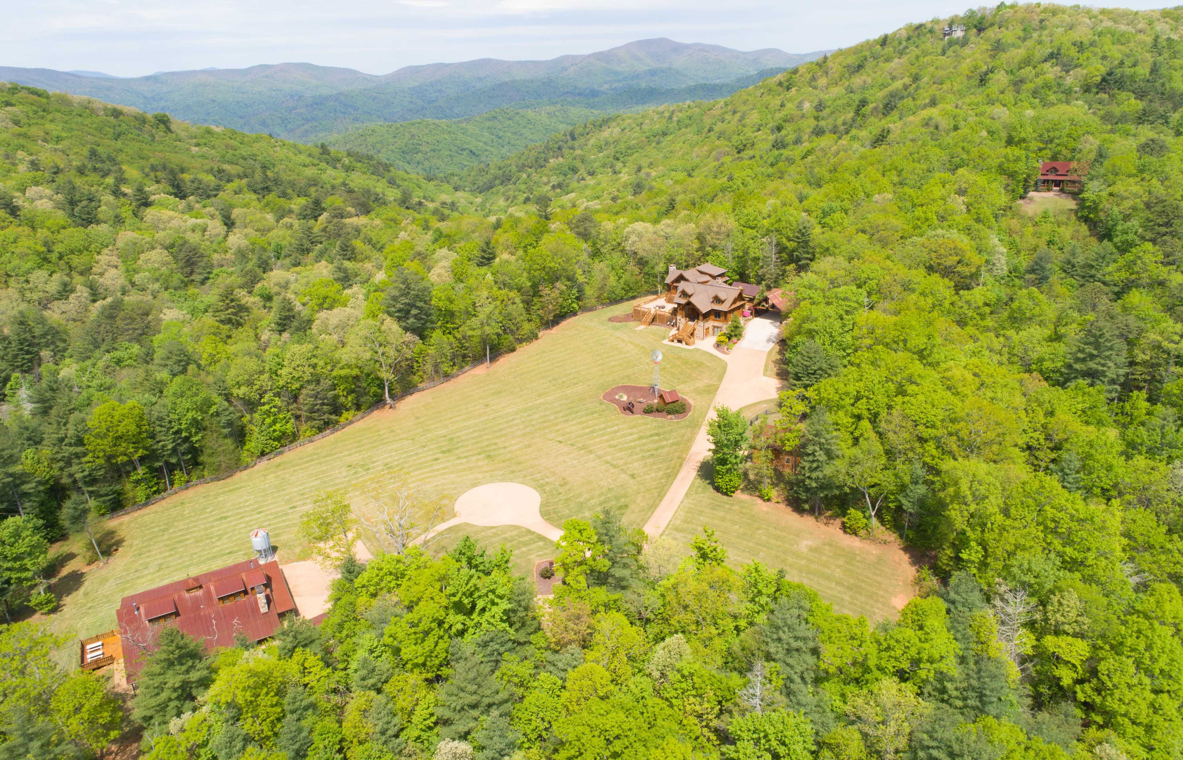 A spacious, green landscape with a large, multi-story house situated on a hillside, surrounded by dense trees and a clear blue sky.