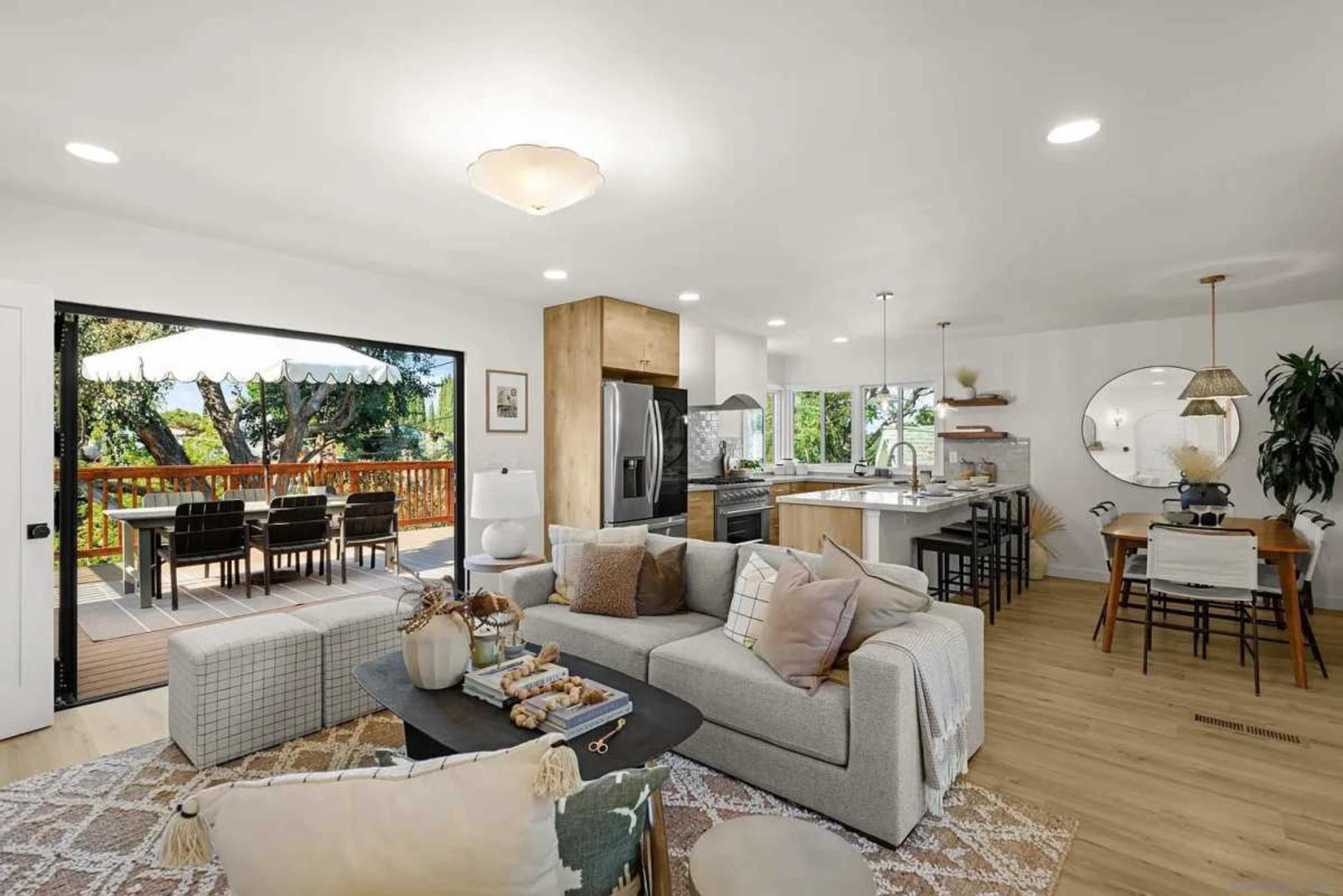 The image shows a modern, open-concept living space featuring a living area with a large sofa, a dining area, and a well-equipped kitchen, all illuminated by natural light from large windows.