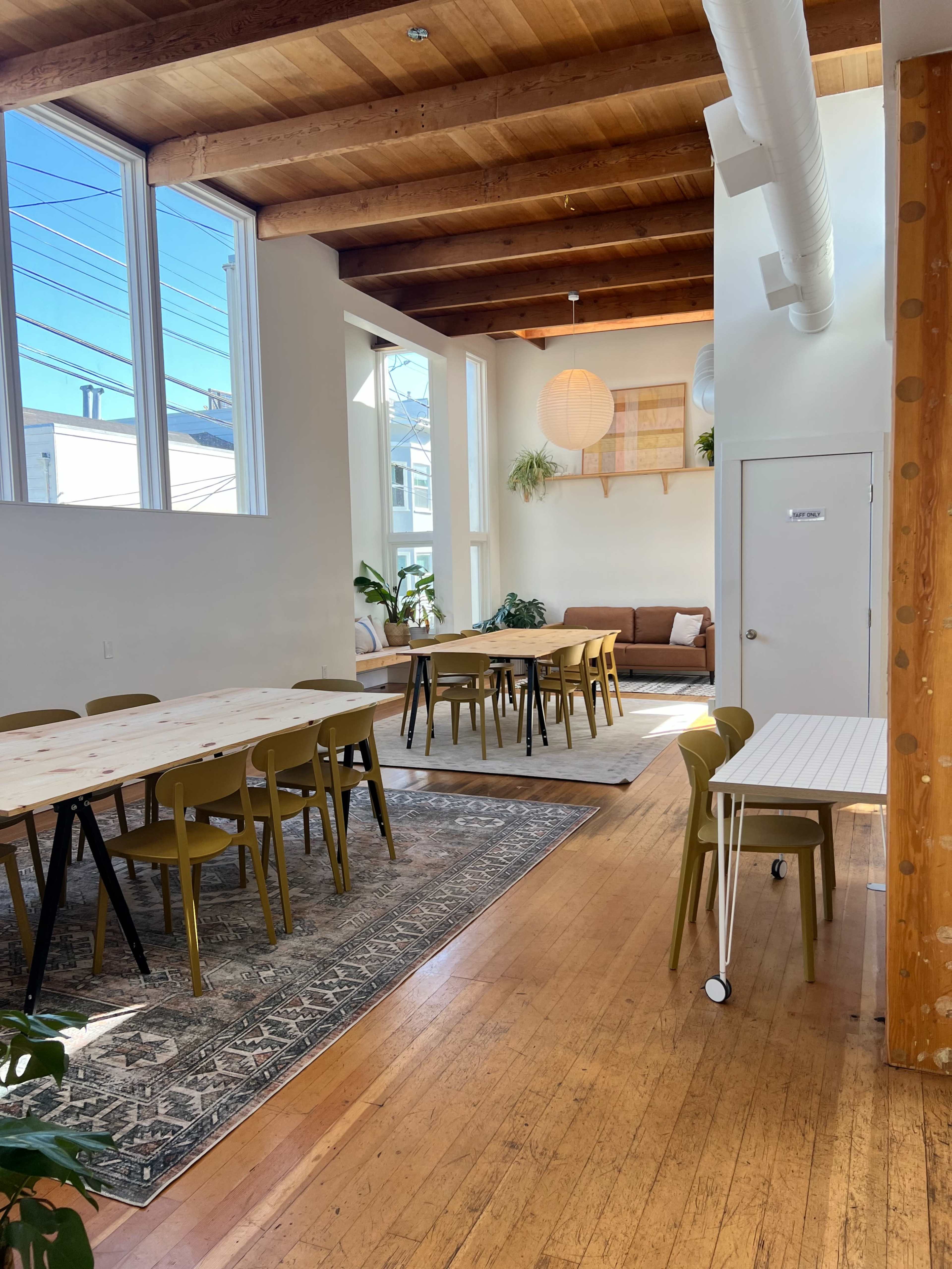 The image shows a bright, airy interior space featuring wooden beams, a mix of tables and chairs, and a cozy seating area with plants and a decorative wall.